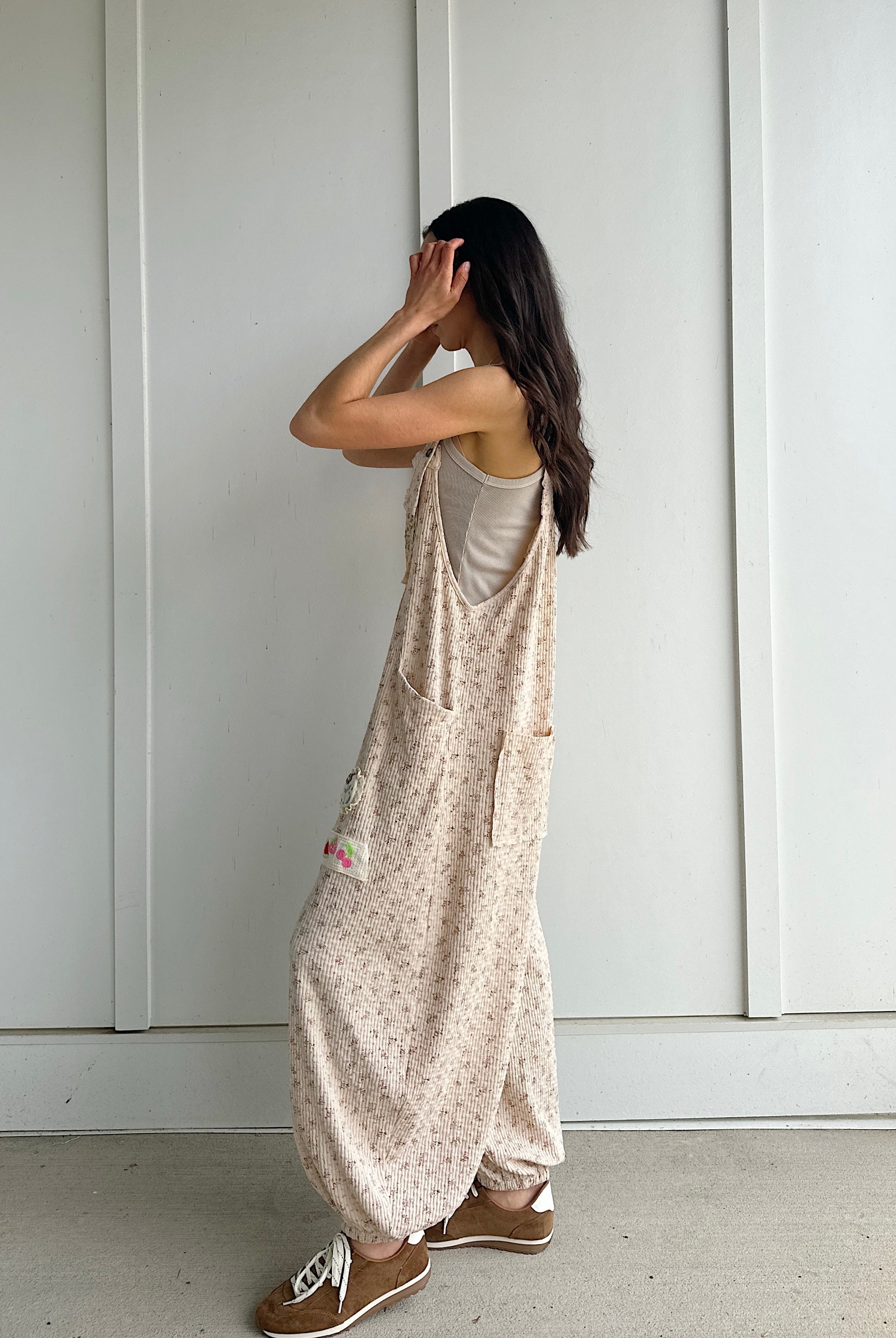 Person wearing a beige patterned overalls standing against a white paneled wall showing side view