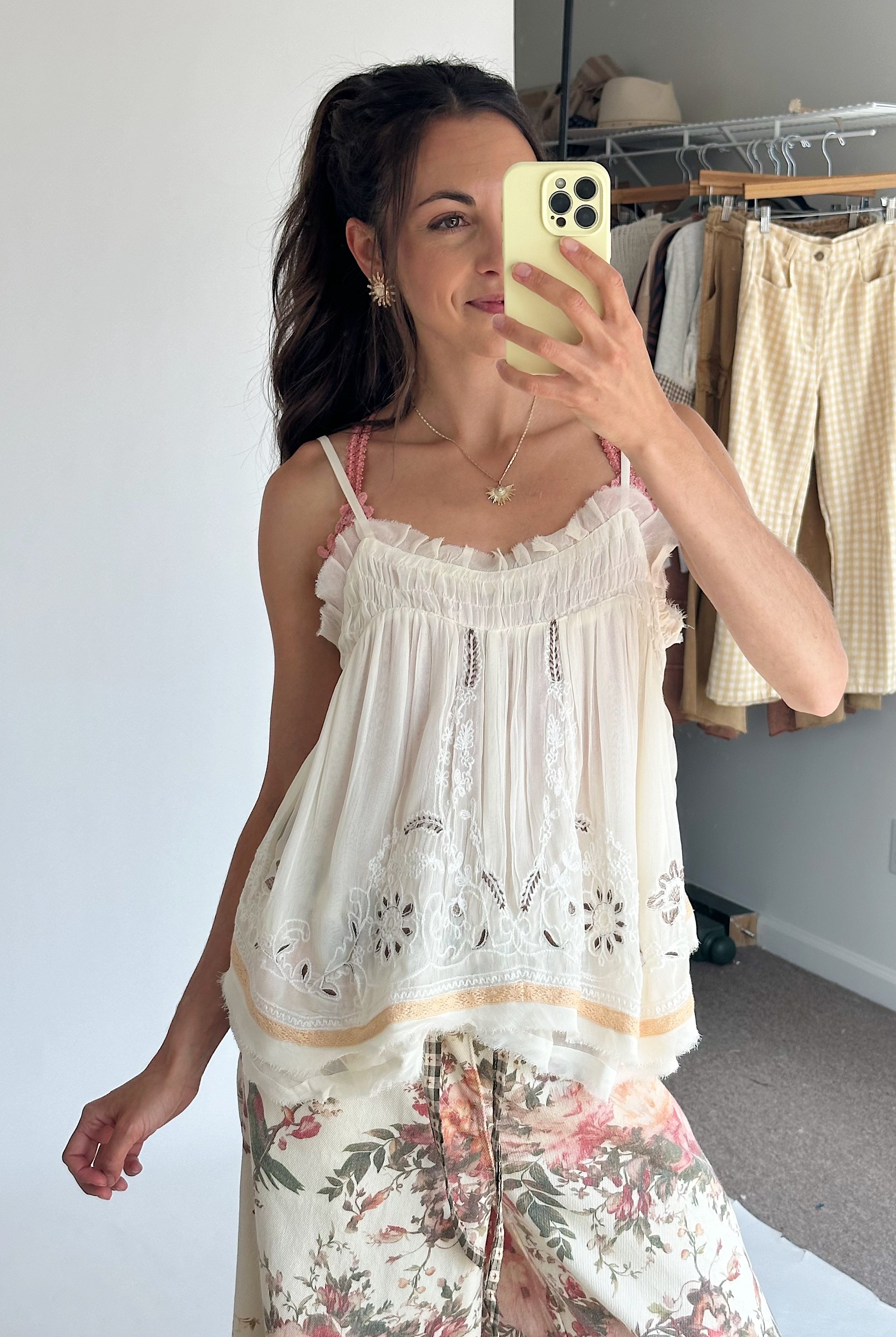 A woman takes a mirror selfie wearing a cream ruffled camisole top with floral embroidery and a floral-printed pants.
