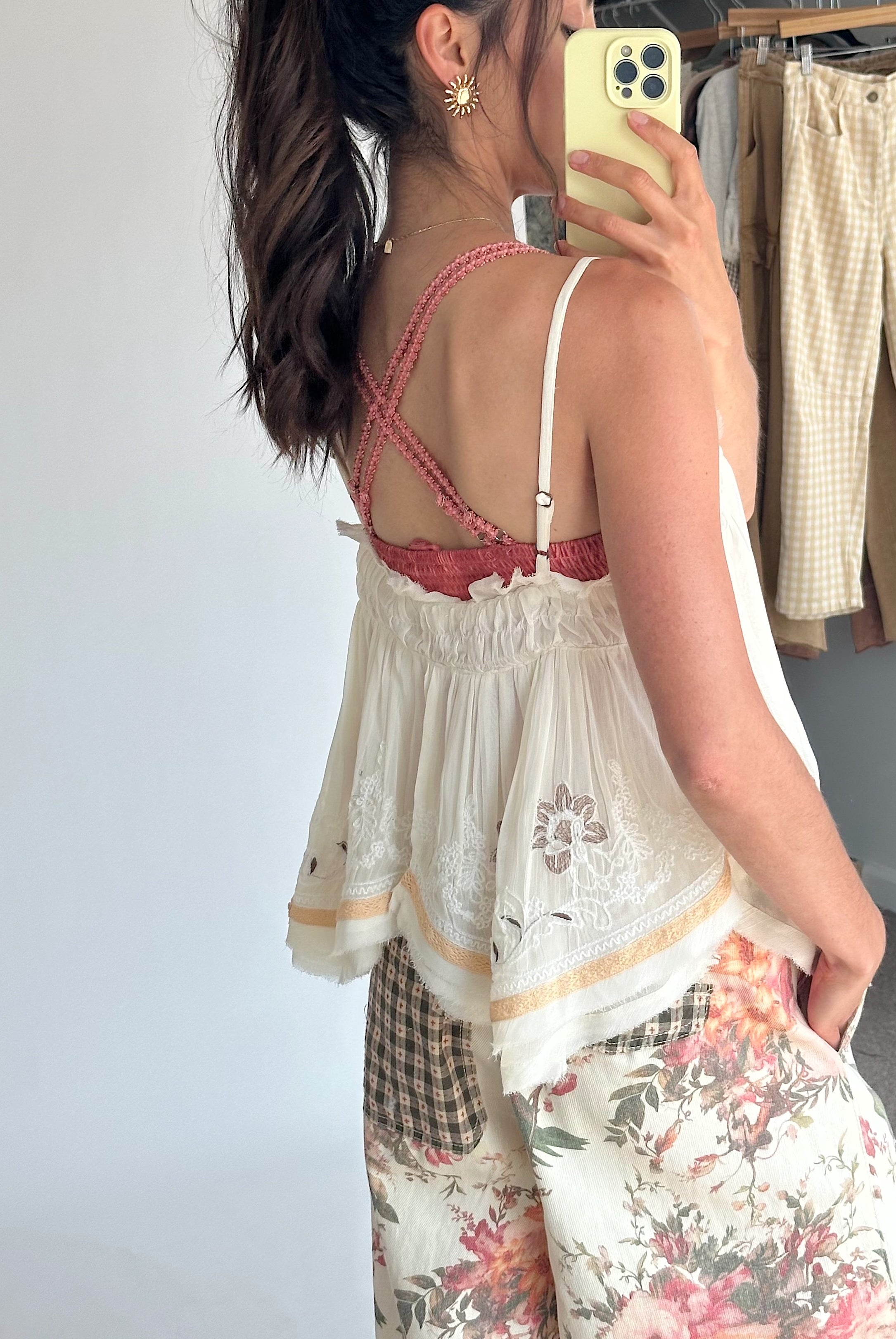 A woman takes a mirror selfie showing the back of a white lace camisole top with a pink braided strap detail and floral wide-leg pants.