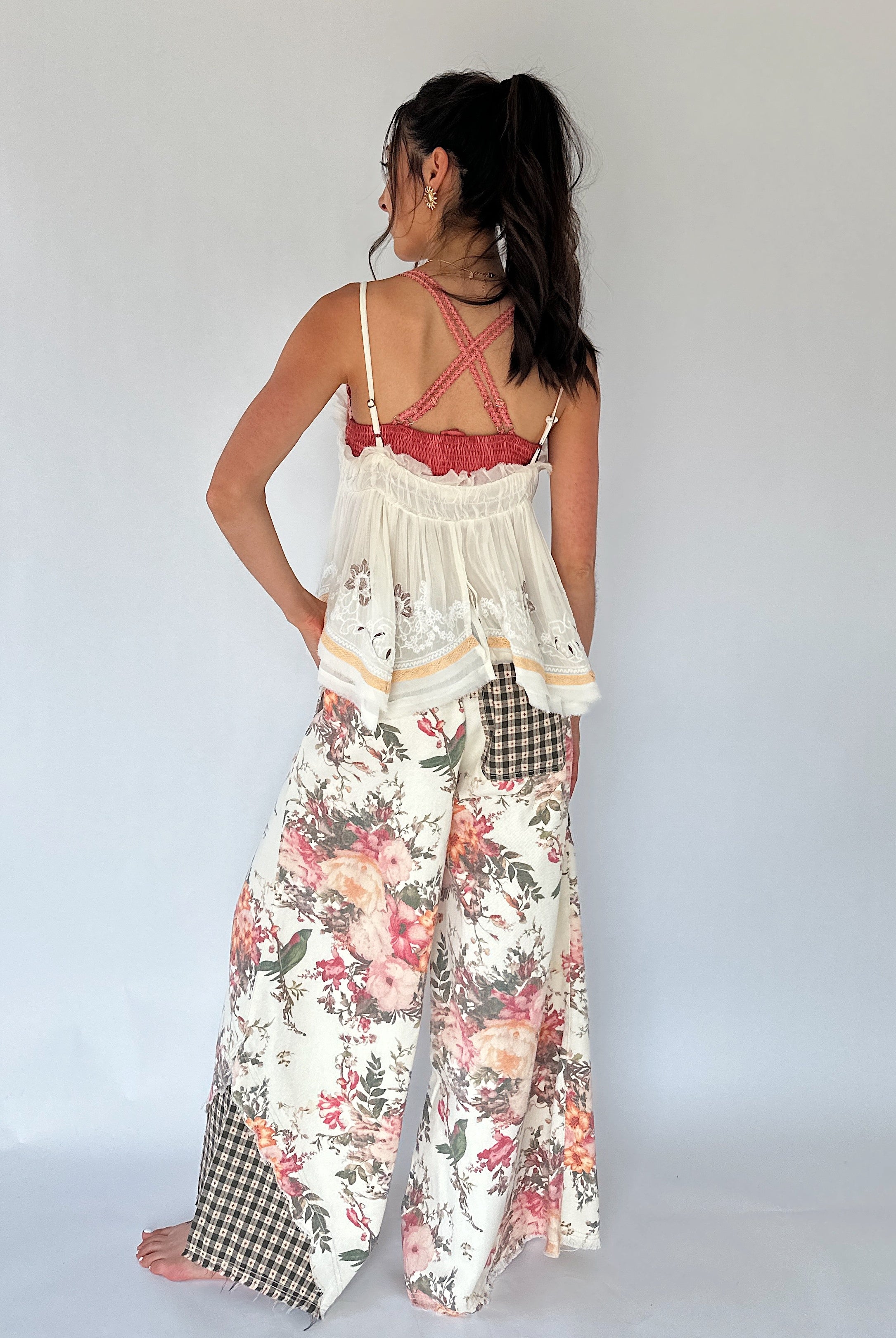 A woman shown from the back wearing a two-piece outfit consisting of a cream lace tank top with thin straps and wide-leg floral pants with gingham patch pockets, set against a plain light gray studio background.