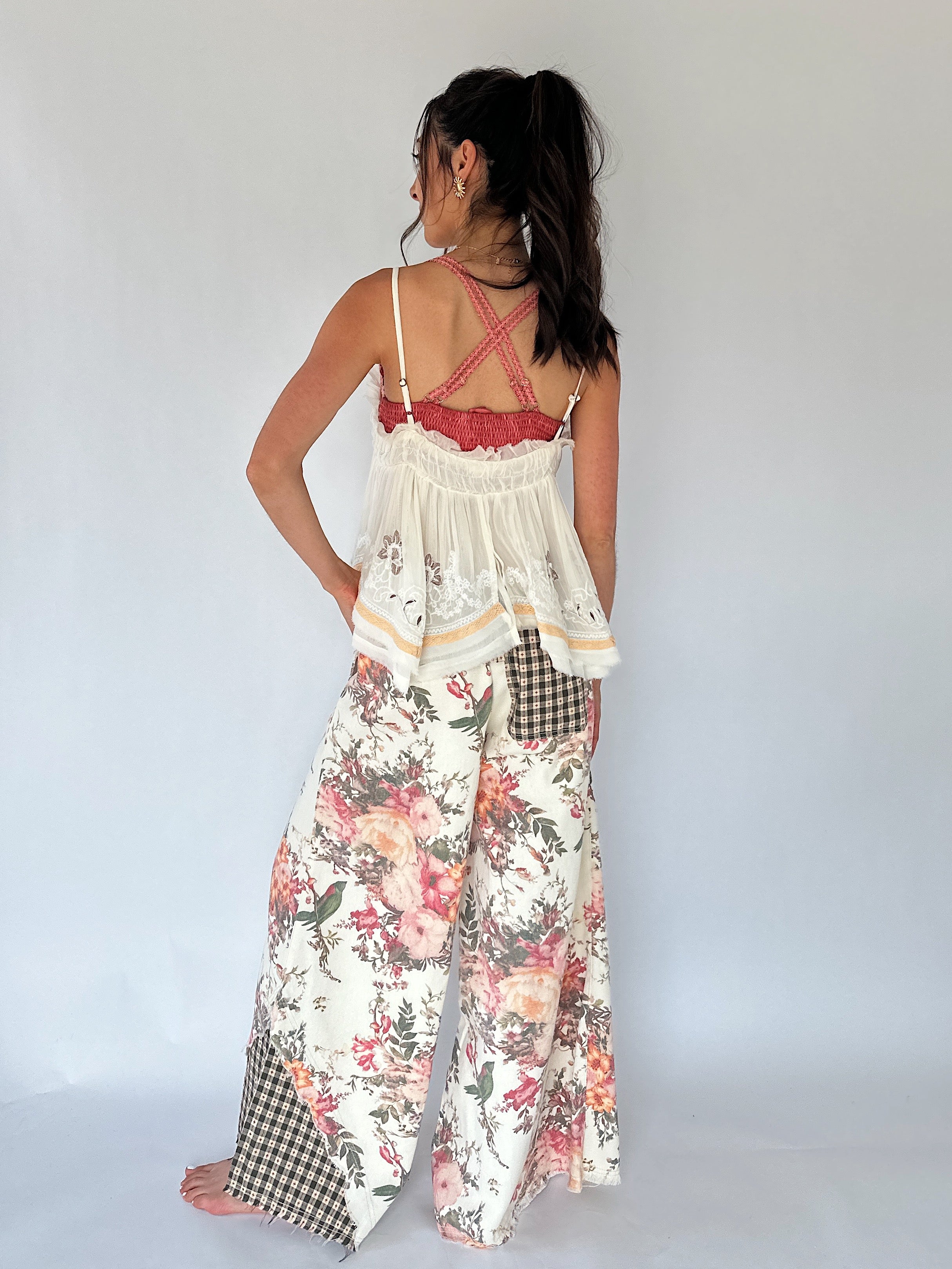 A woman shown from the back wearing a two-piece outfit consisting of a cream lace tank top with thin straps and wide-leg floral pants with gingham patch pockets, set against a plain light gray studio background.