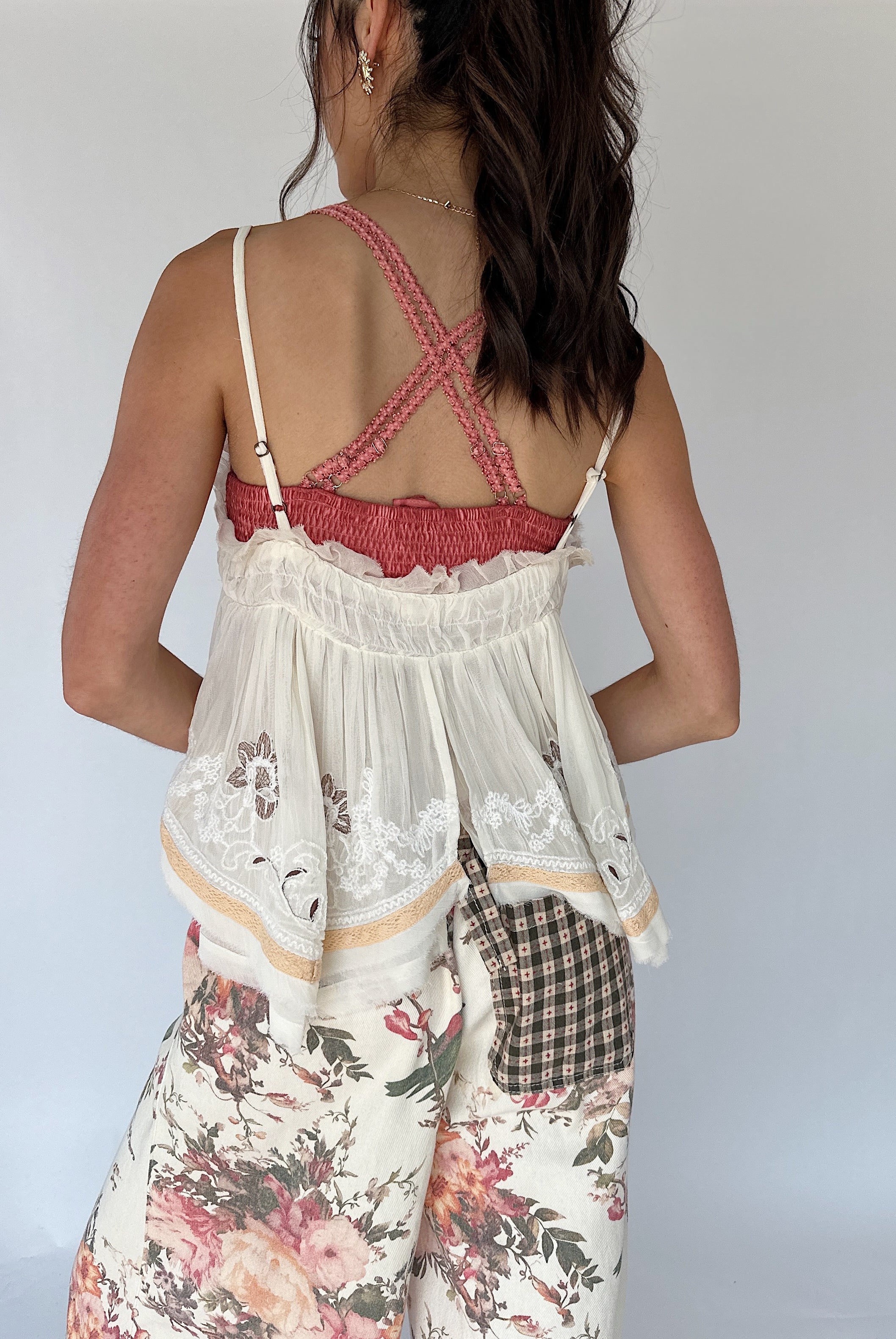 A woman shown from the back wearing a white lace-trim camisole top with a pink ruffled criss-cross strap design and floral print pants against a plain light gray background.