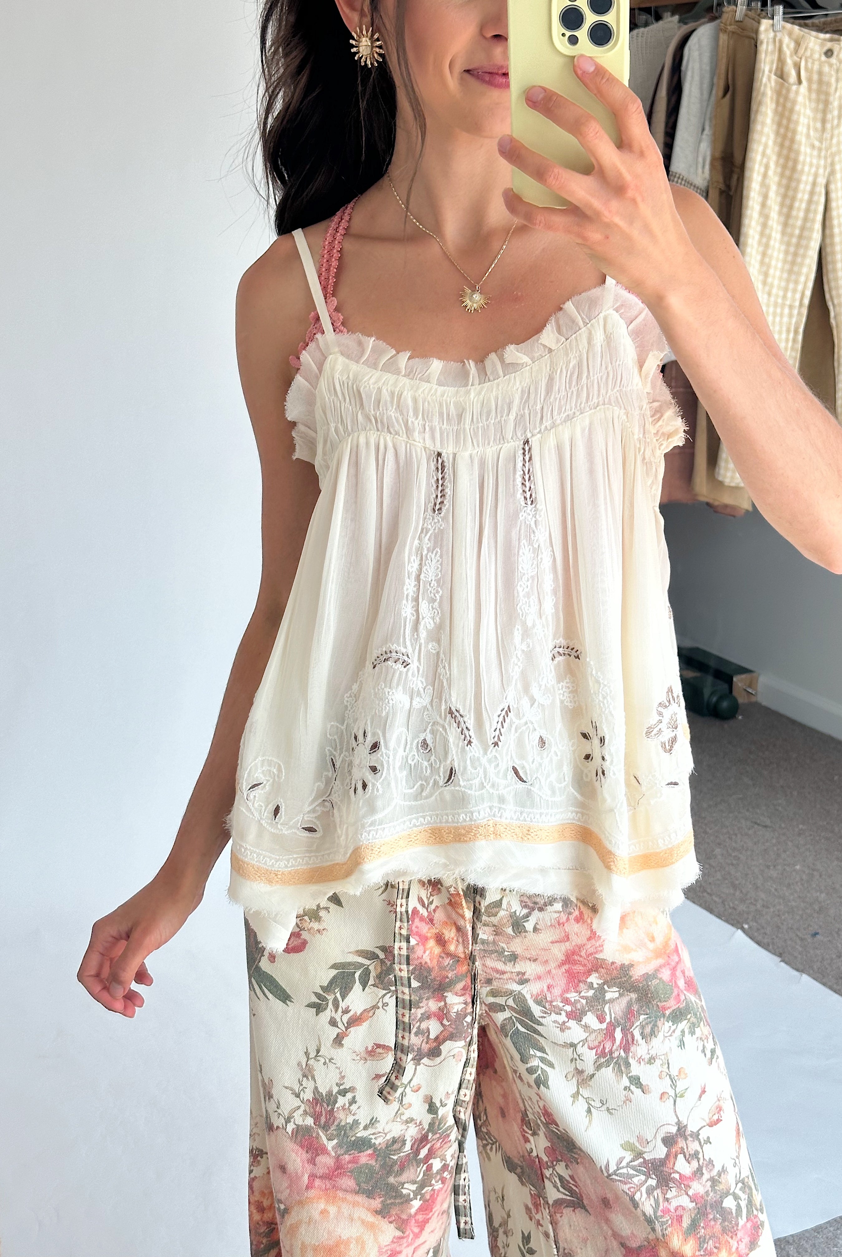 A woman takes a mirror selfie wearing a white ruffled camisole top with delicate embroidery and a floral print pants featuring pink, red, and green botanical patterns. She is holding a light yellow smartphone in front of her face.
