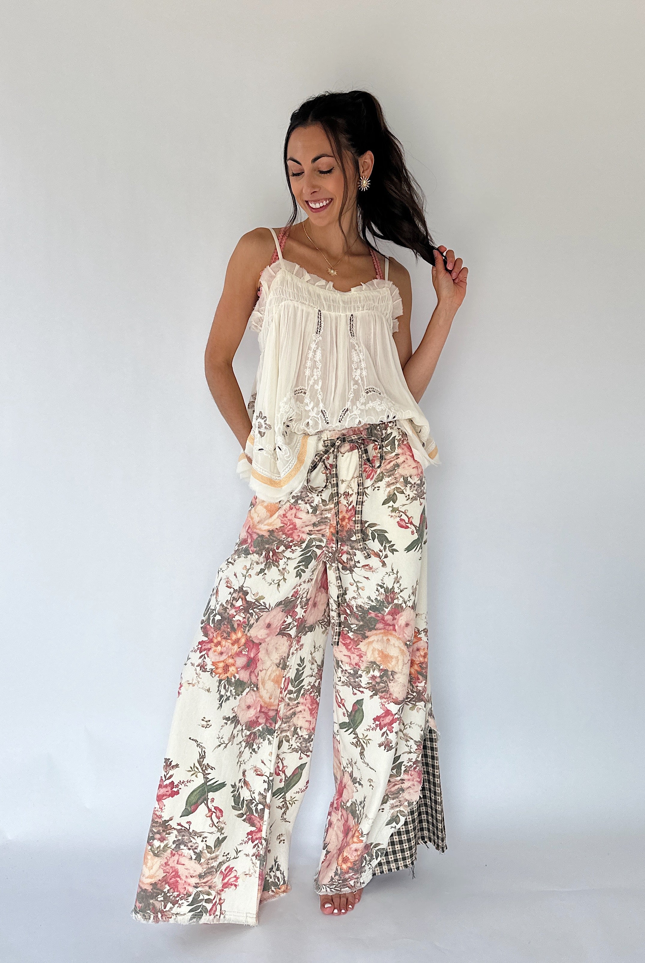 A woman modeling a bohemian-style two-piece outfit featuring a cream lace-trimmed camisole top and wide-leg pants with a pink, red, and green floral print, set against a plain white studio background.