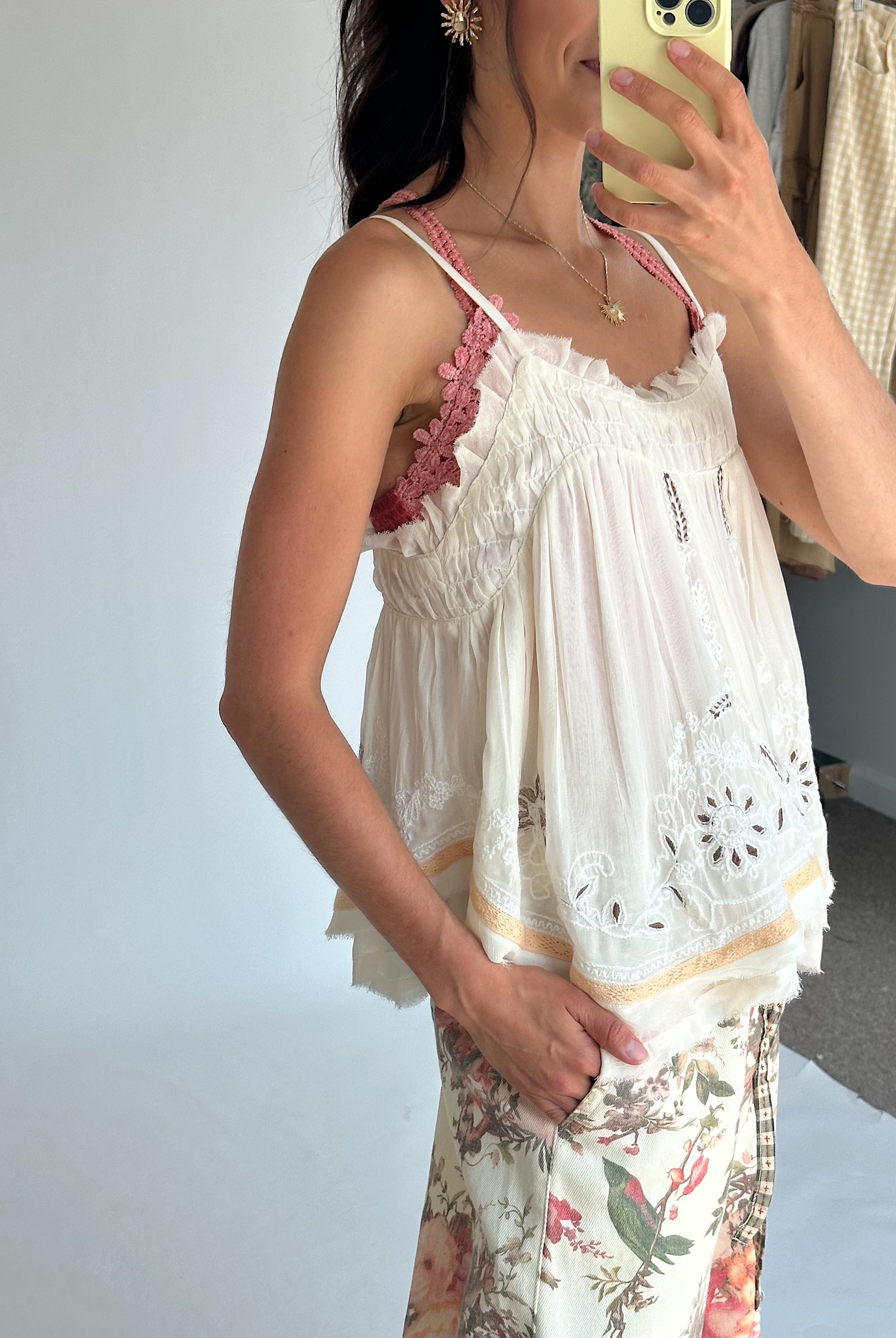 A woman takes a mirror selfie wearing a white eyelet lace camisole top over a pink bralette and floral print pants. She is holding a light yellow smartphone.