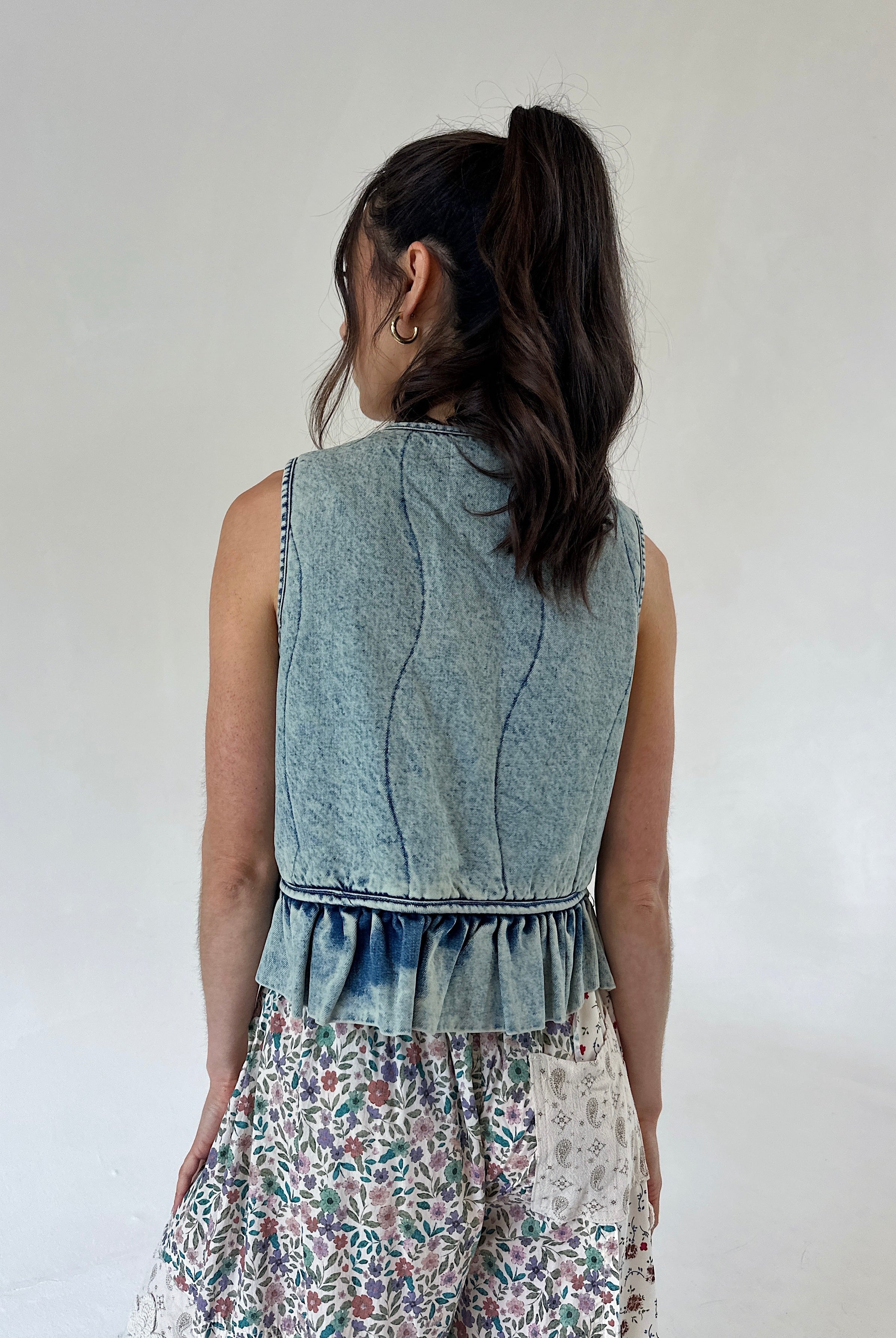 A woman shown from the back wearing a sleeveless light blue denim vest with a ruffled hem with floral pants, set against a plain off-white background.