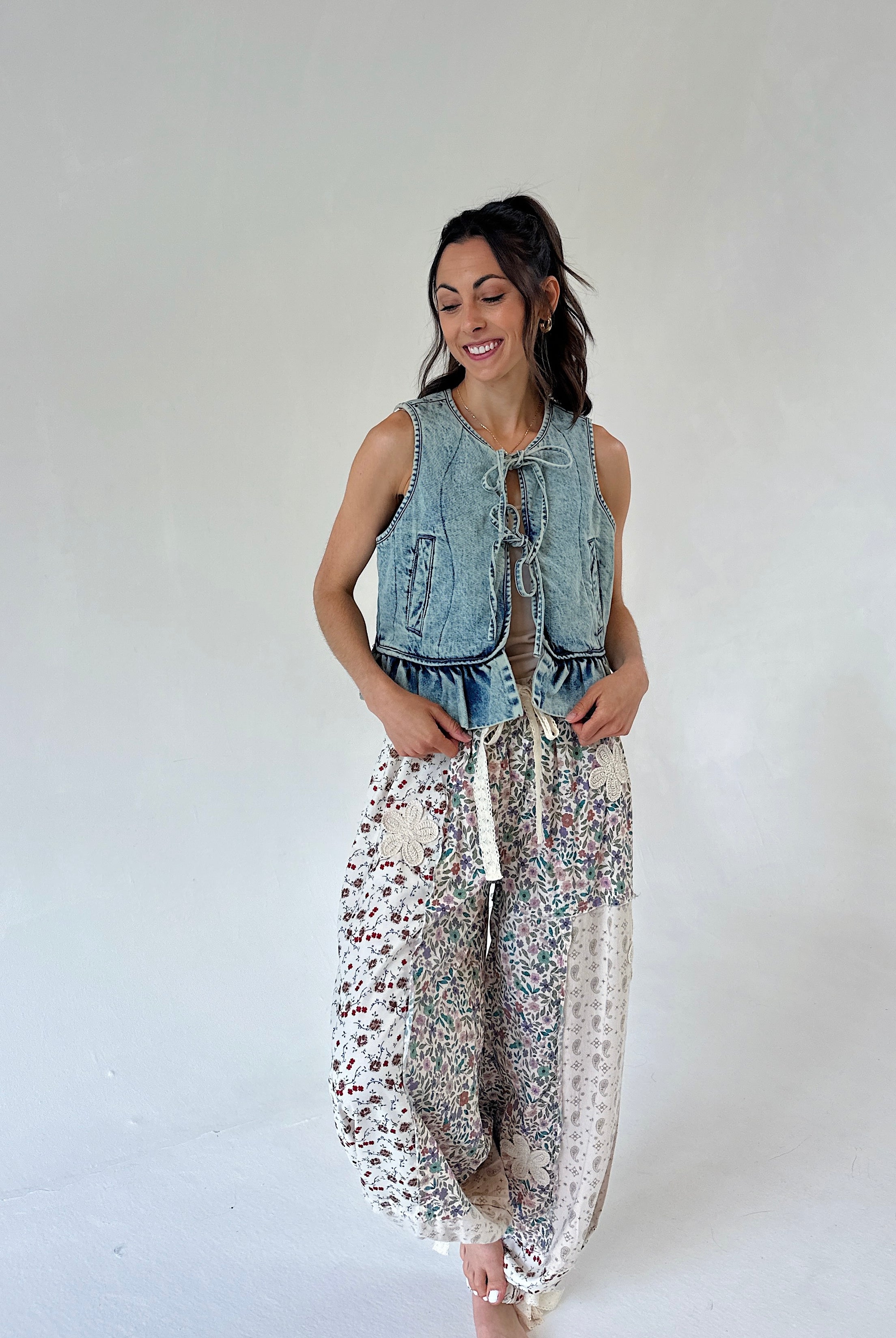 A woman modeling a sleeveless light-wash denim vest with front tie details and loose white harem-style pants covered in a small multicolor floral print, against a plain light gray studio background.