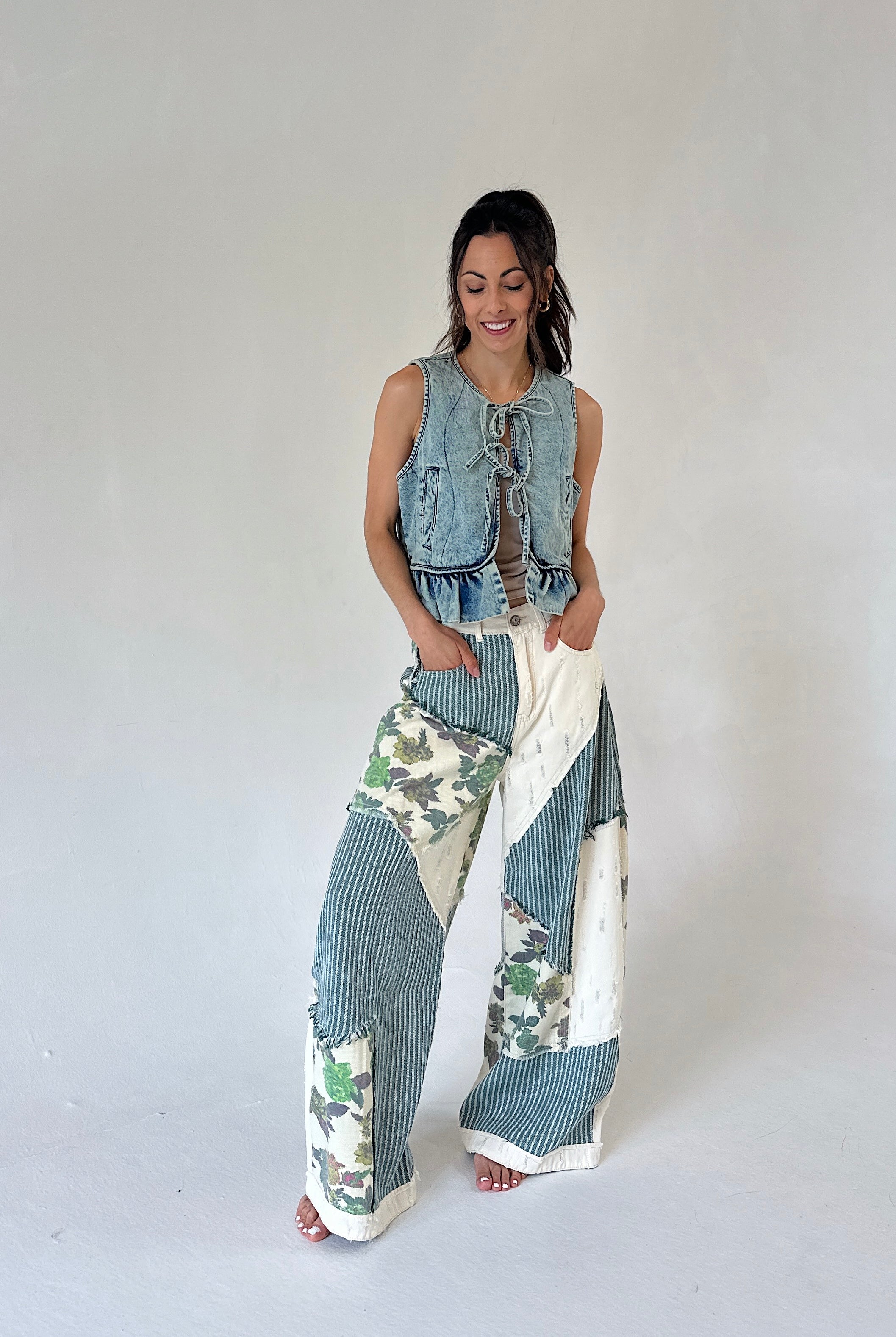 A woman modeling a sleeveless light blue denim vest paired with wide-leg patchwork pants featuring blue-gray and white stripes, floral prints, and solid white sections, against a plain light gray studio background.