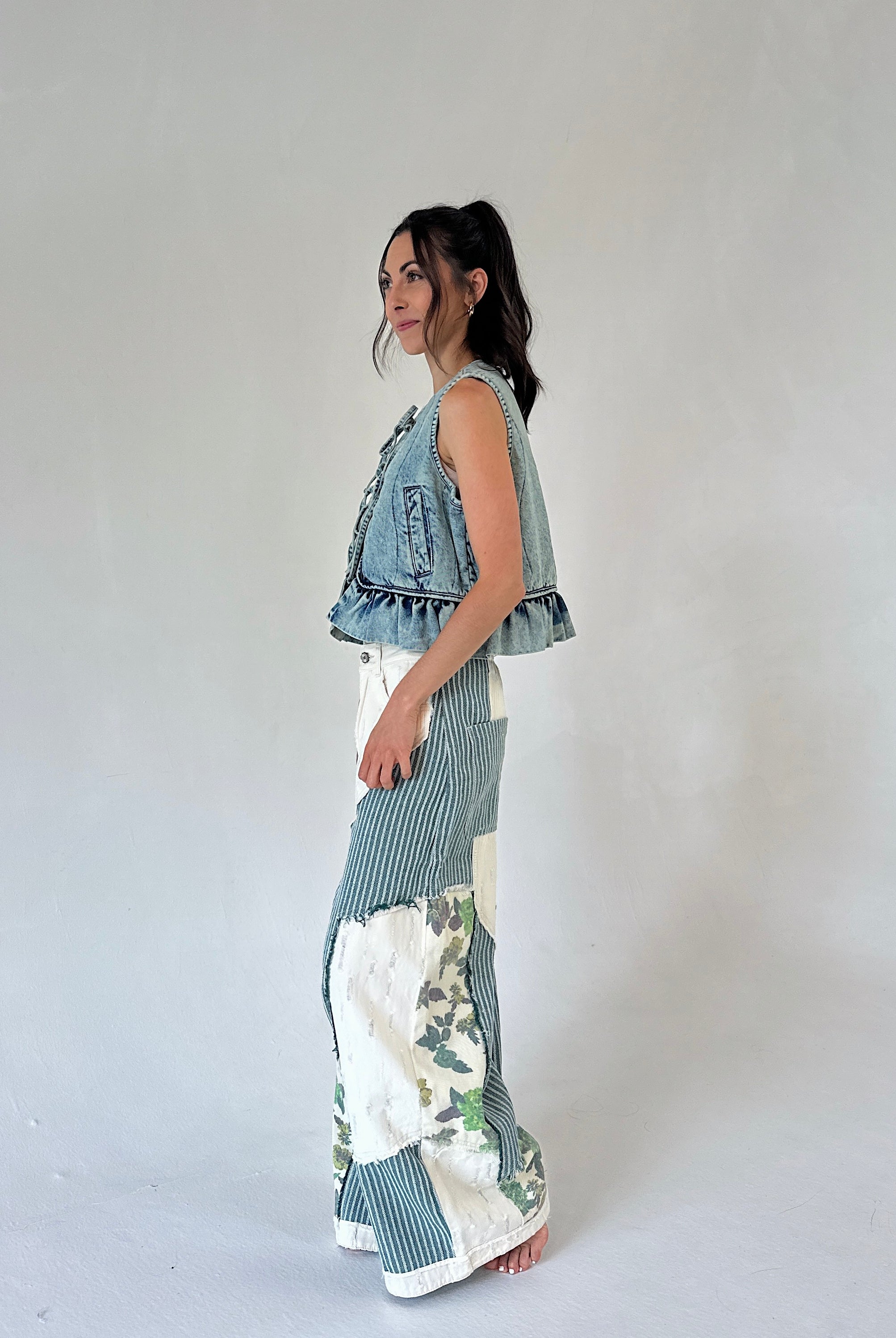 A woman stands in profile wearing a sleeveless denim vest with ruffled trim and wide-leg patchwork pants featuring blue stripes and white floral sections, against a plain light gray studio background.
