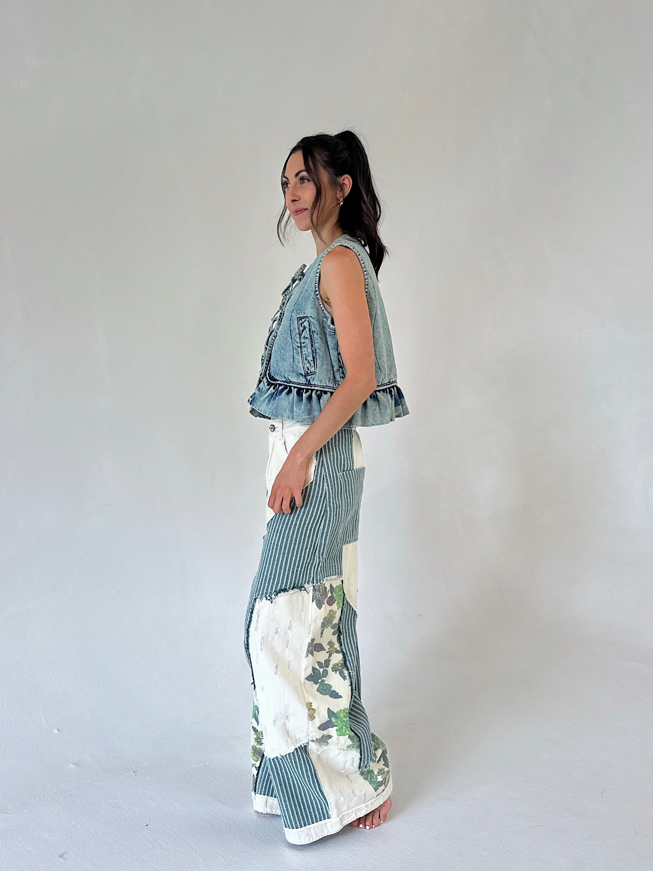 A woman stands in profile wearing a sleeveless denim vest with ruffled trim and wide-leg patchwork pants featuring blue stripes and white floral sections, against a plain light gray studio background.