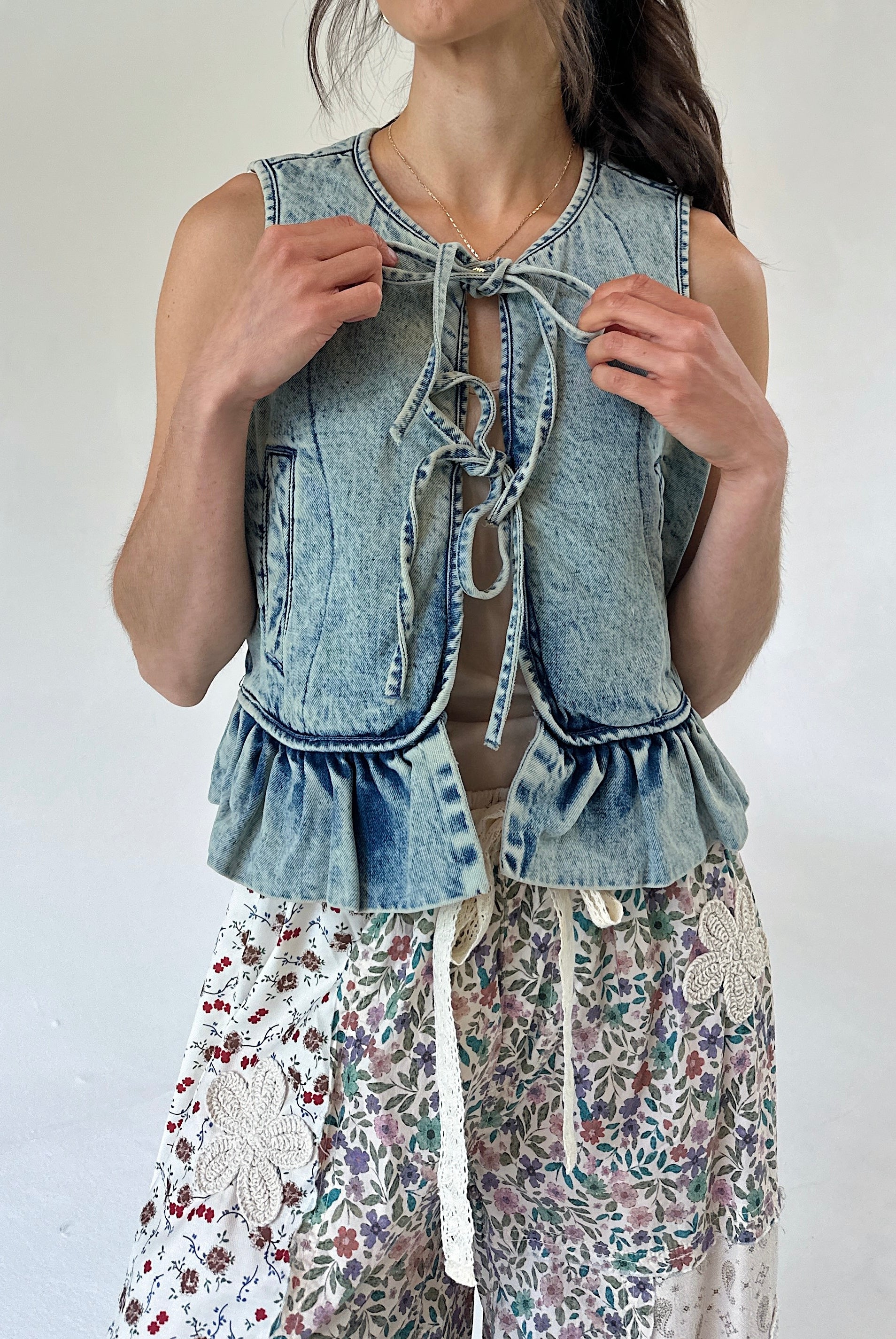 A woman wearing a sleeveless light-wash denim vest with tie-front detailing and ruffled hem, paired with floral print pants, against a plain light background.