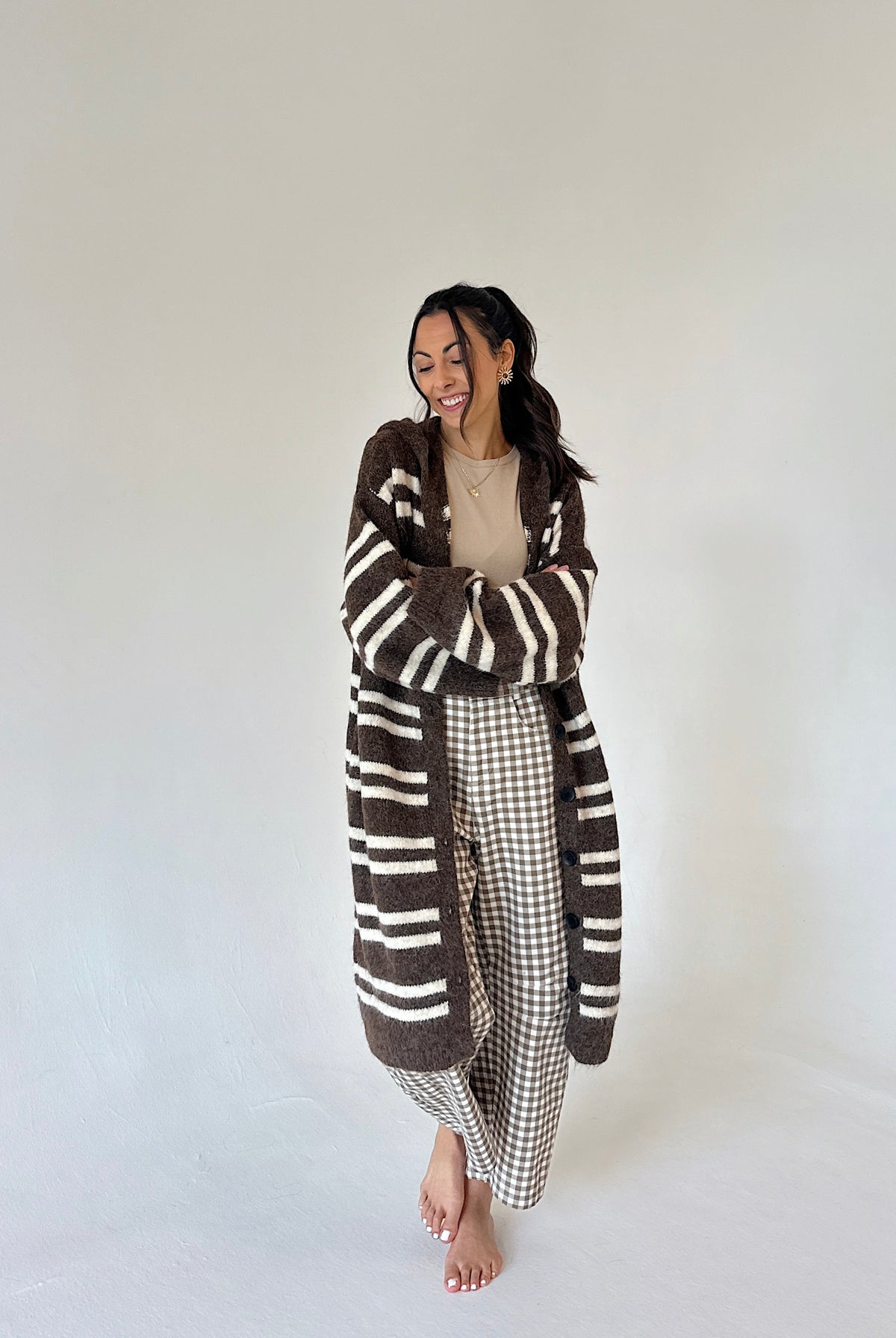 A woman stands barefoot against a plain off-white studio background, wearing a long brown and cream striped fuzzy cardigan over a beige top and brown-and-white gingham pants.