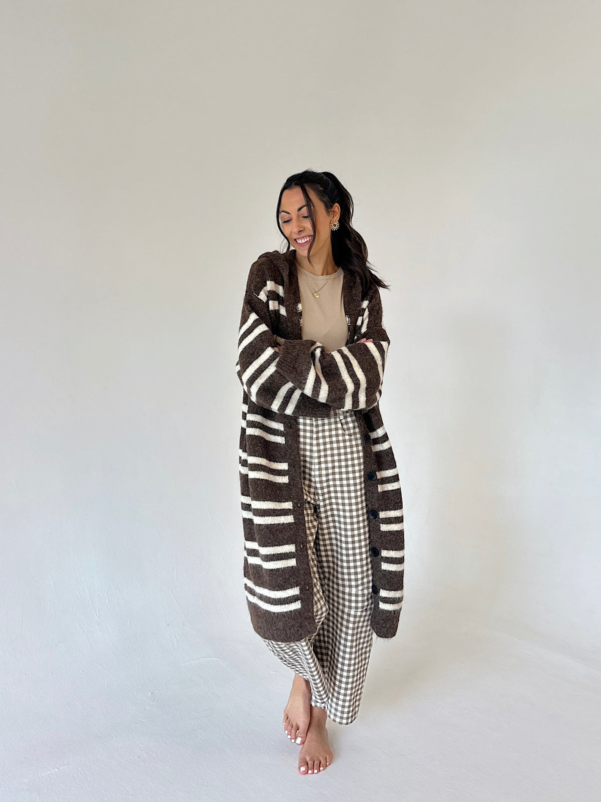 A woman stands barefoot against a plain off-white studio background, wearing a long brown and cream striped fuzzy cardigan over a beige top and brown-and-white gingham pants.