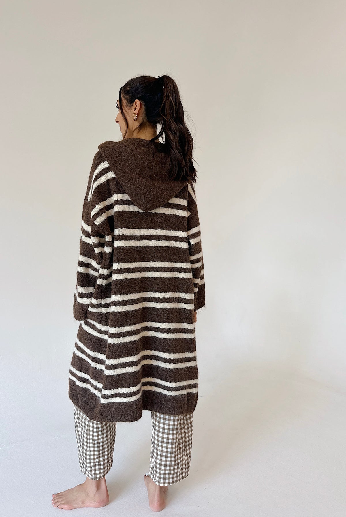 A woman stands barefoot against a plain off-white wall, viewed from the back, wearing a brown and cream horizontally striped hooded cardigan and brown-and-white checkered pajama pants.