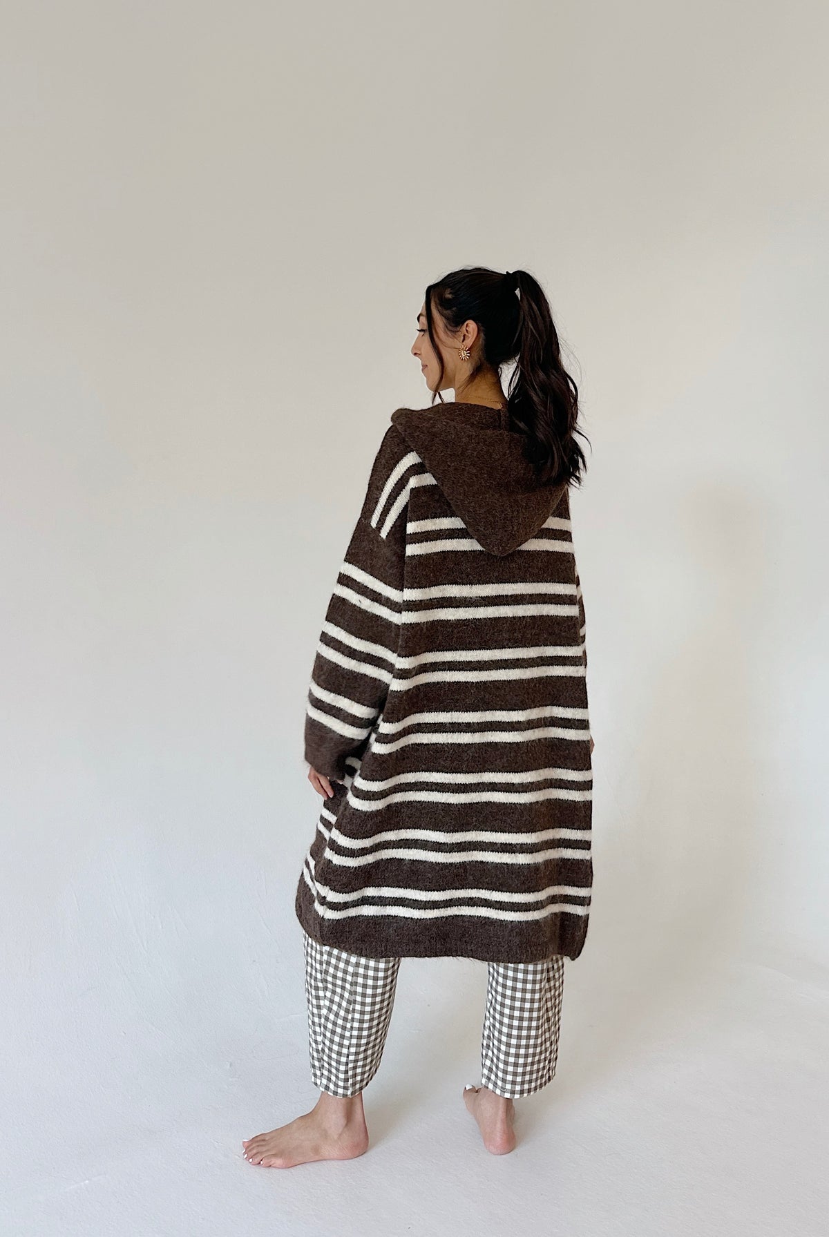 A woman stands barefoot in a studio wearing a brown and cream striped hooded cardigan or poncho-style top, paired with gingham pants, photographed against a plain off-white background.