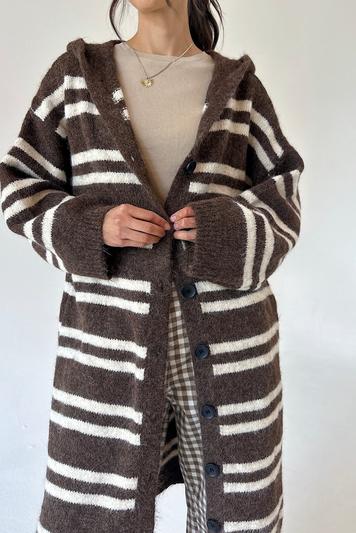 A person wearing a brown and cream striped fuzzy knit cardigan with a hood and front buttons, layered over a beige top, against a plain light background.