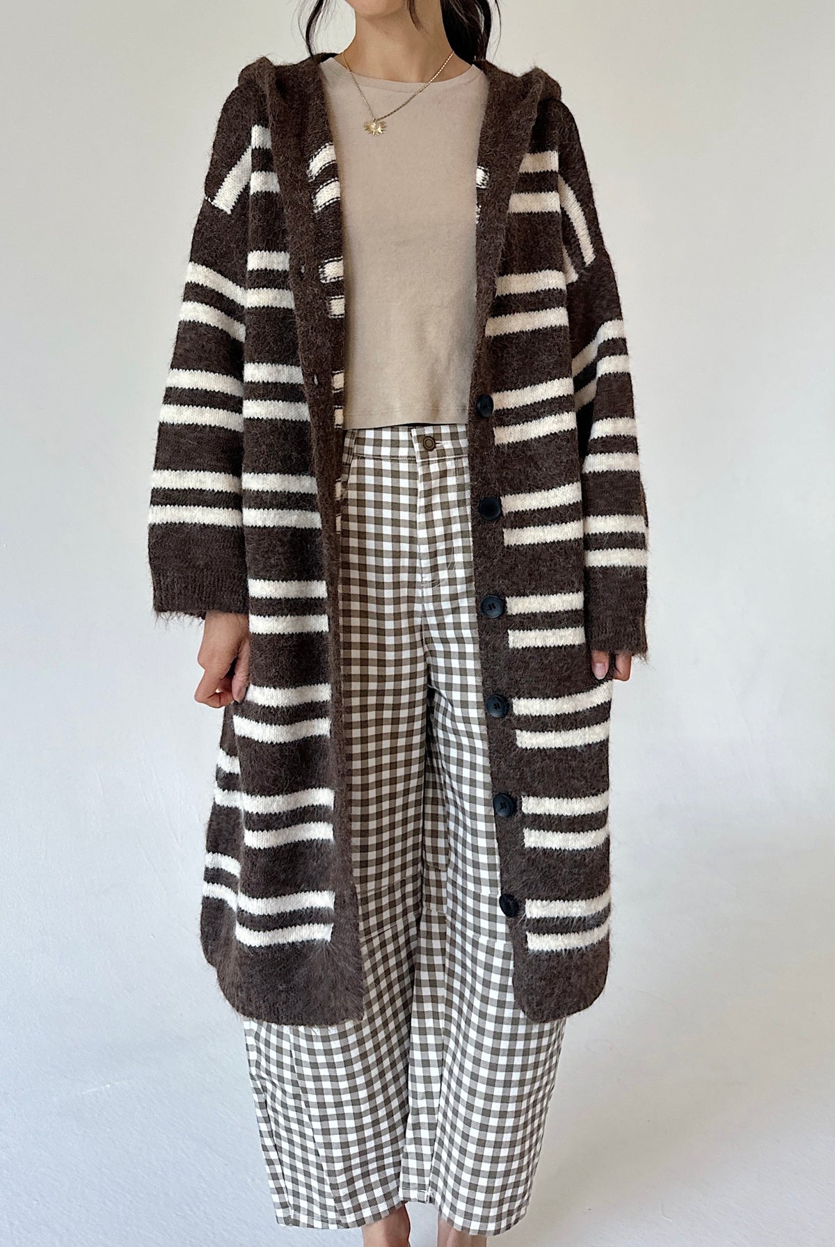 A person wearing a long brown and cream striped fuzzy cardigan over a beige top and brown-and-white gingham cropped pants, standing against a plain light background.