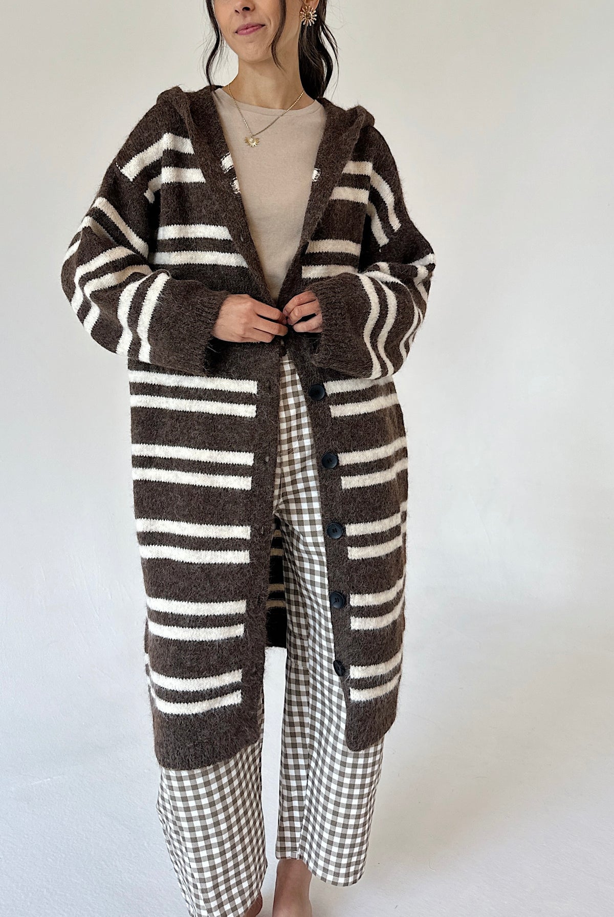 A woman wearing a long brown and cream striped knit cardigan over a beige top and brown-and-white gingham pants, standing against a plain light background.