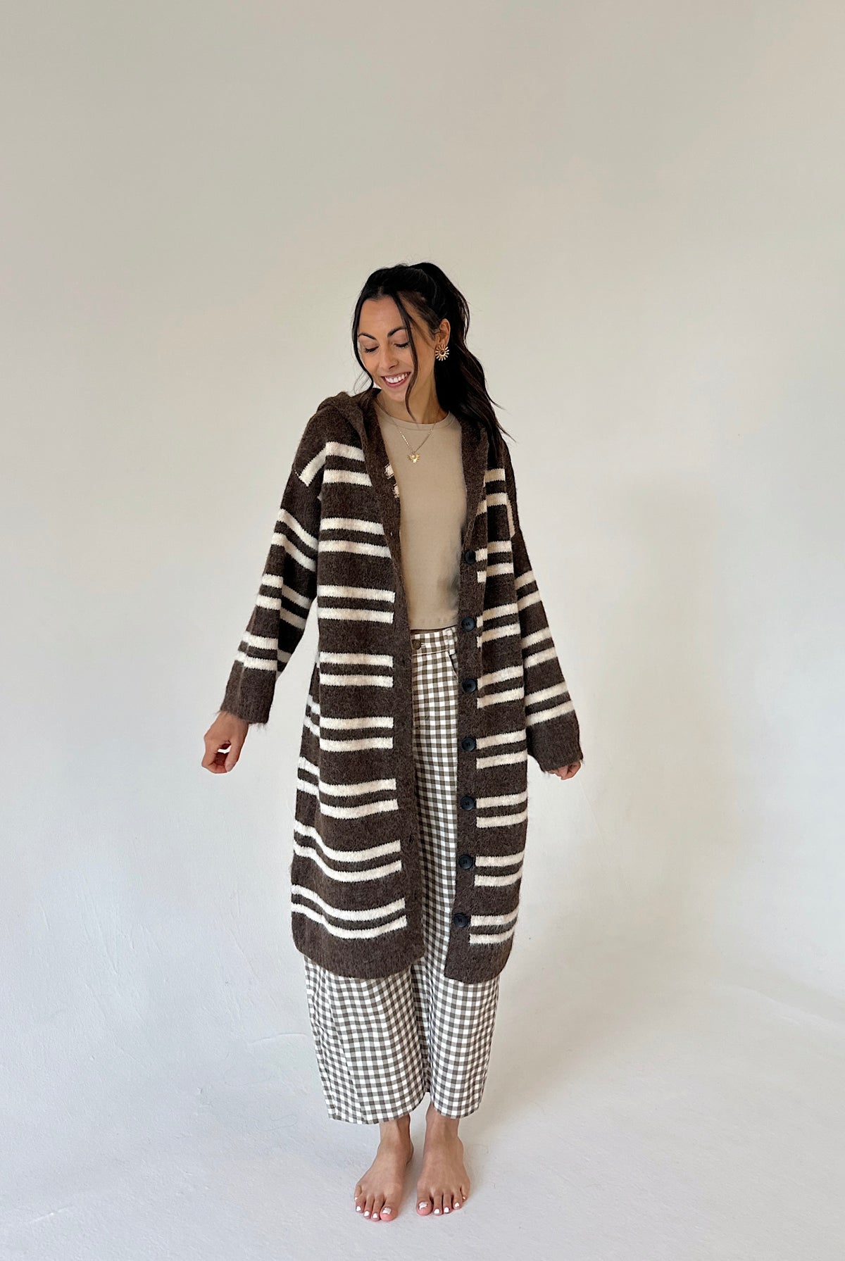 A woman stands barefoot against a plain light background wearing a long brown and cream striped fuzzy cardigan over a beige top and brown-and-white gingham cropped pants.