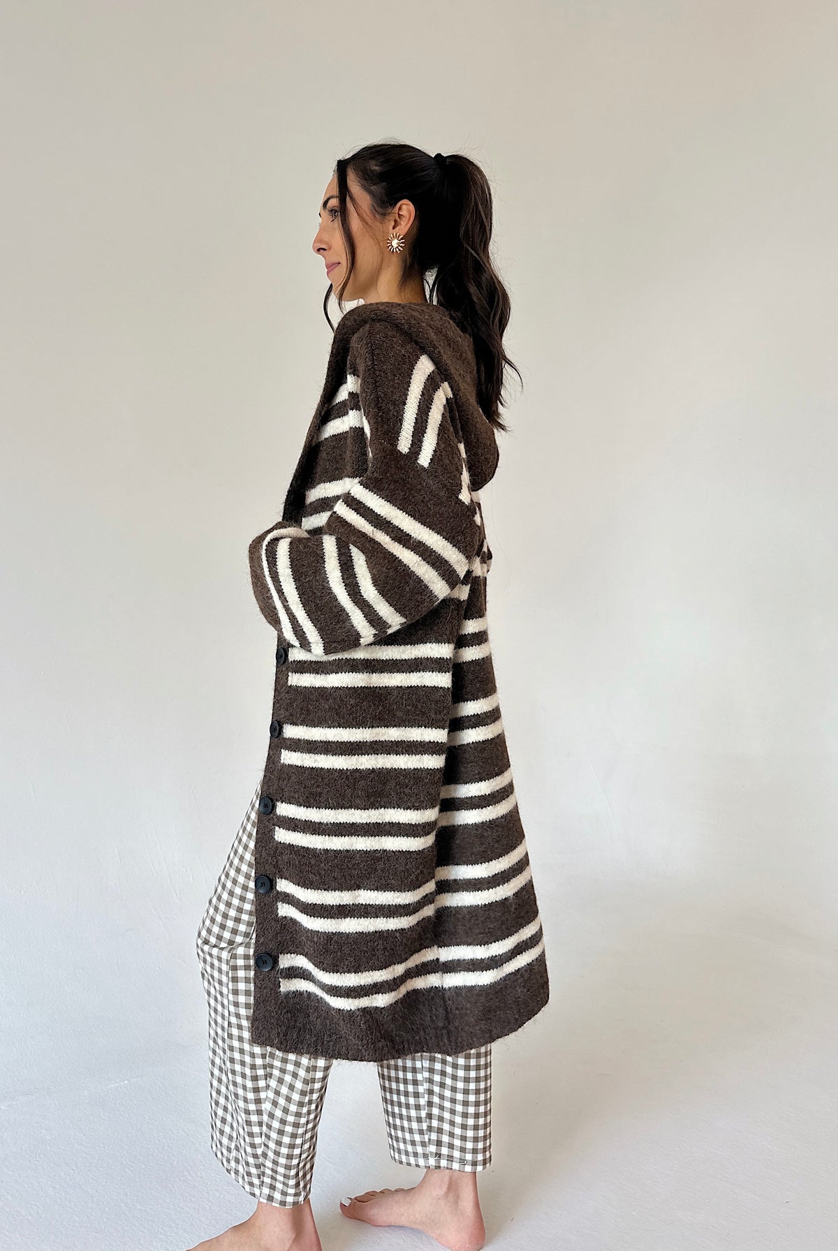 A woman stands barefoot in profile wearing a long brown and cream striped fuzzy cardigan over a brown-and-white gingham outfit, photographed against a plain light gray studio background.