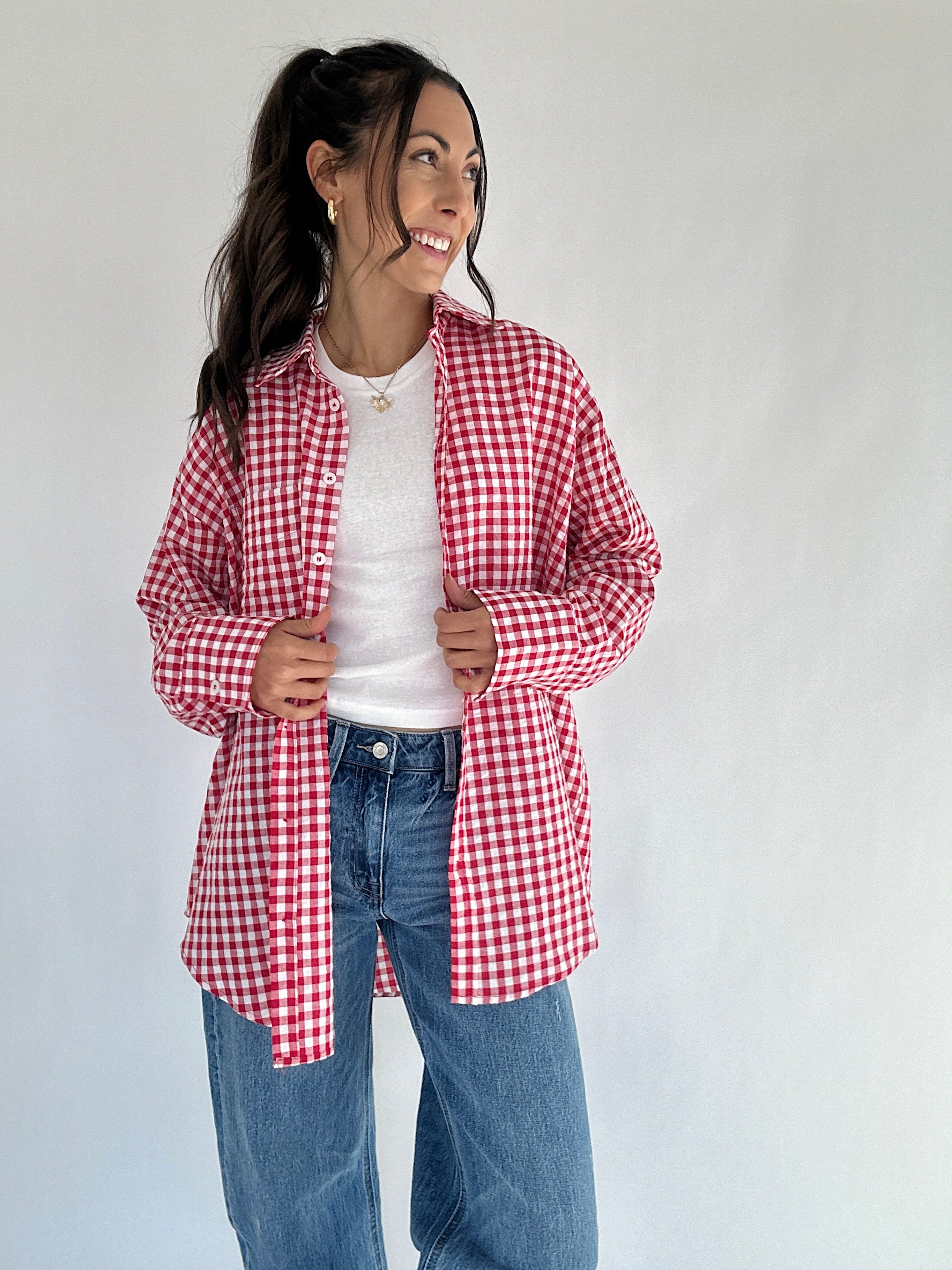 Woman wearing a red and white checkered shirt over a white top and blue jeans on a plain background