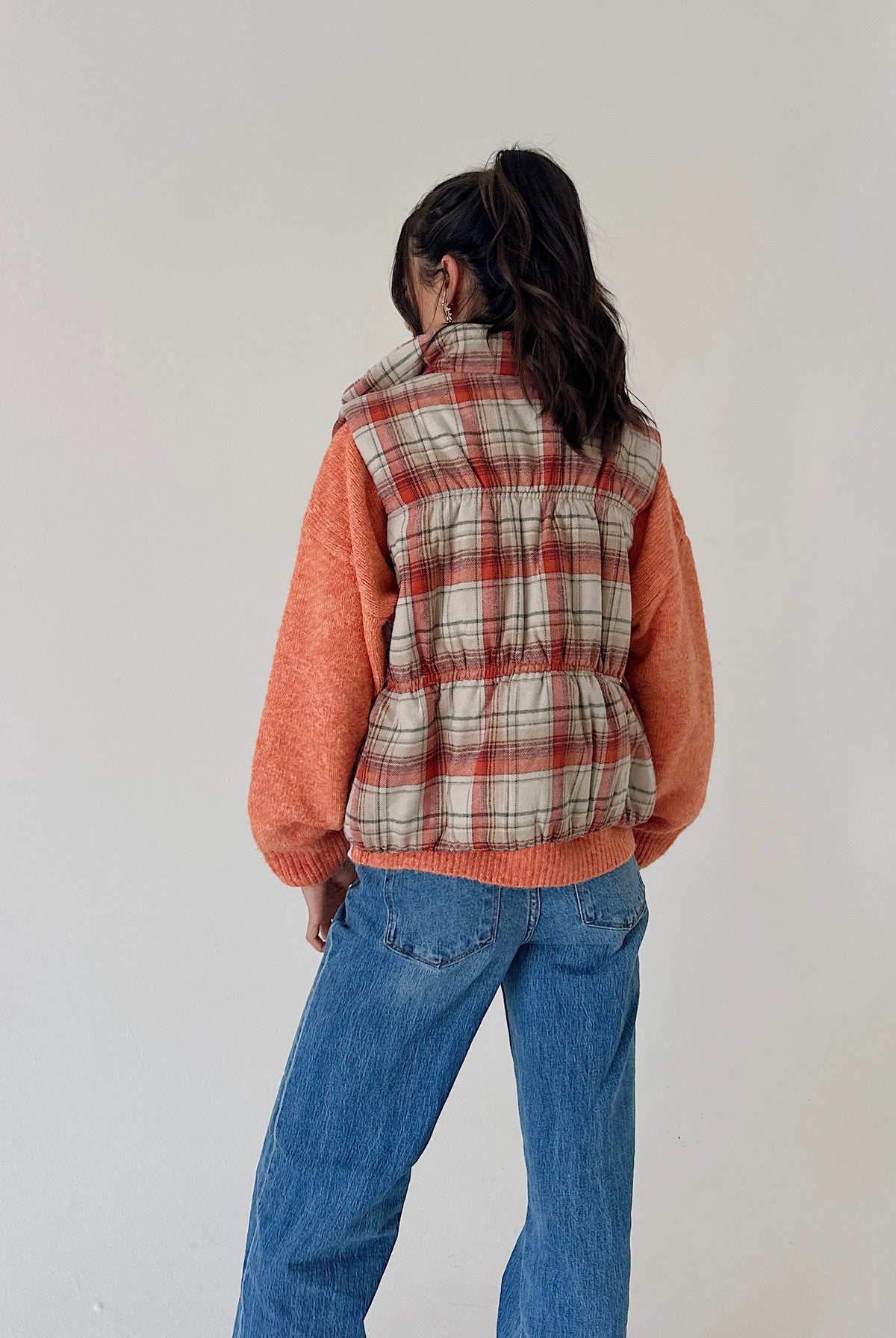 A person shown from the back wearing a sleeveless puffer vest with a beige, red, and white plaid pattern over a fuzzy orange sweater, paired with blue jeans, against a plain light gray wall.