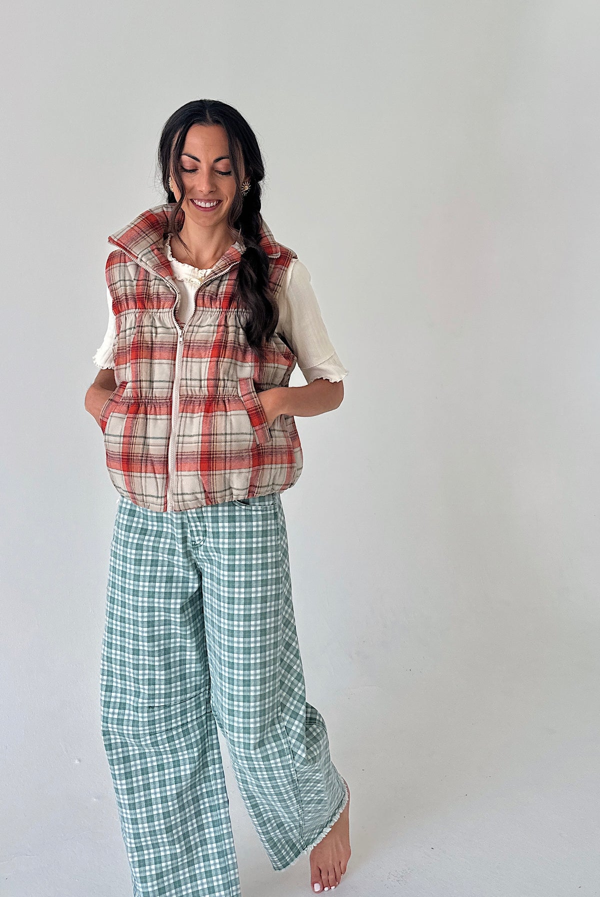 A woman modeling a plaid sleeveless puffer vest over a white ruffled shirt and green plaid wide-leg pants, standing against a plain light gray studio background.