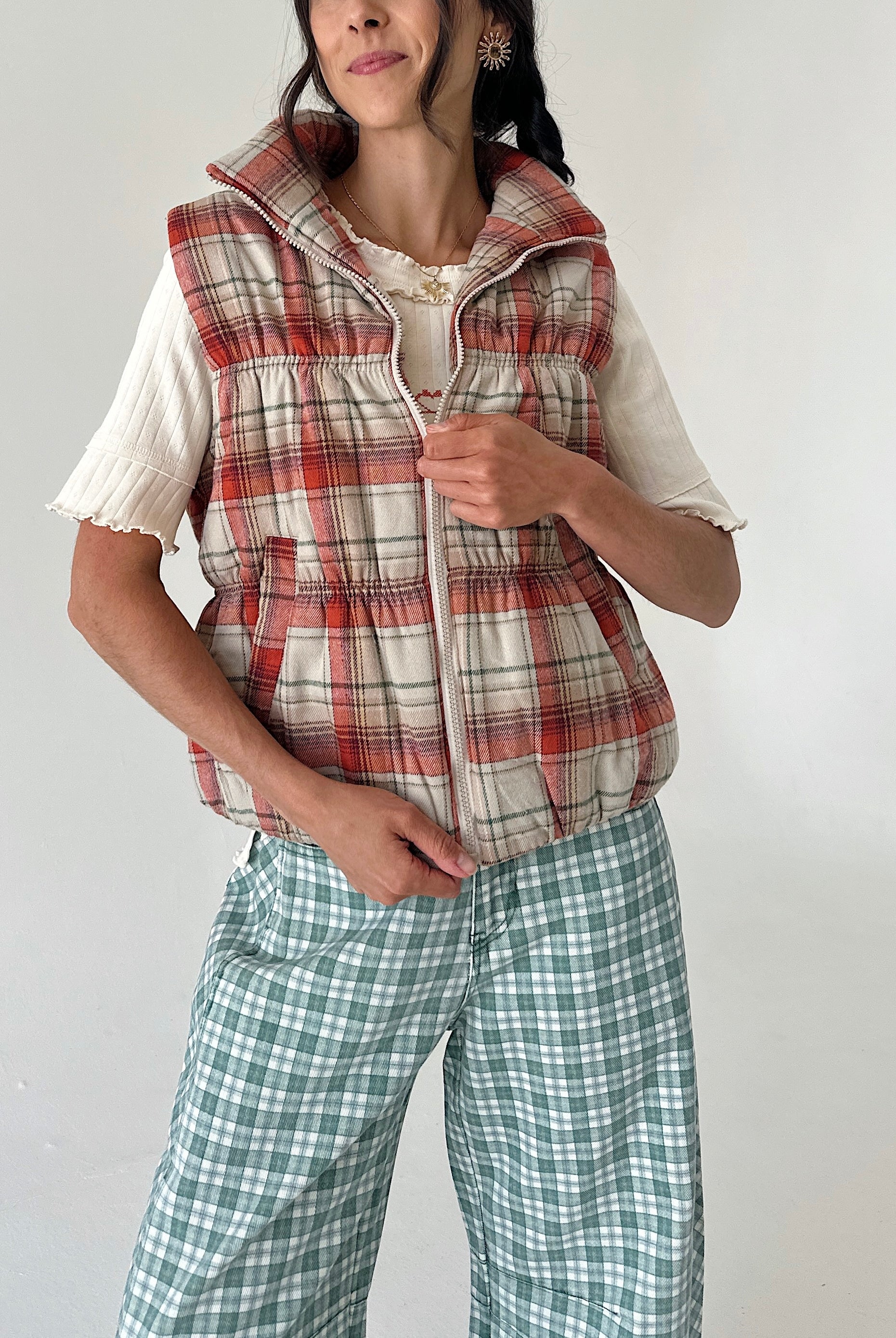 A woman wearing a sleeveless zip-front puffer vest in a red, beige, and white plaid pattern over a cream short-sleeve top, paired with green and white plaid pants, against a plain light gray background.