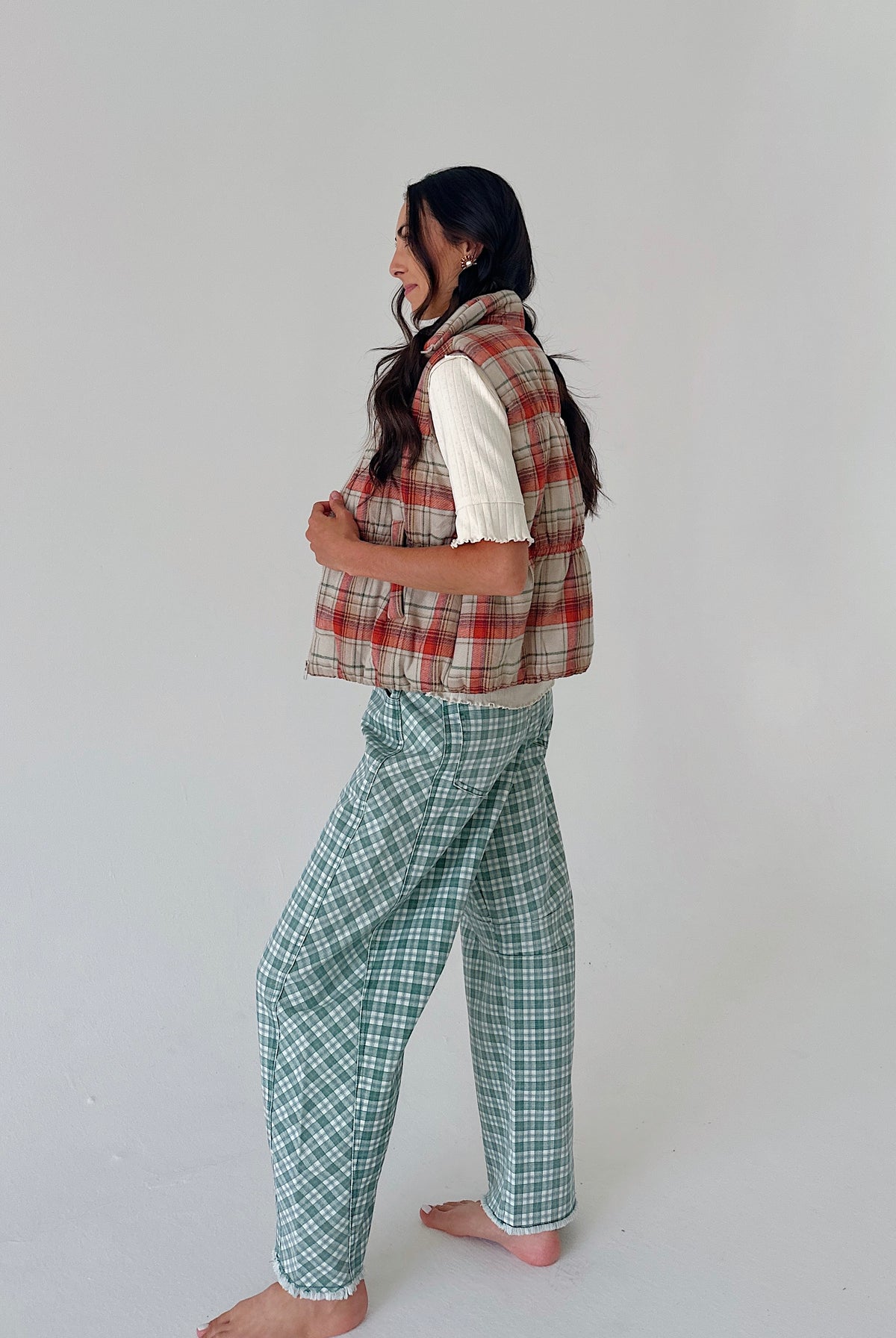 A woman stands barefoot in profile wearing a sleeveless red and beige plaid vest over a white short-sleeve top and green-and-white plaid pants, against a plain light gray studio background.