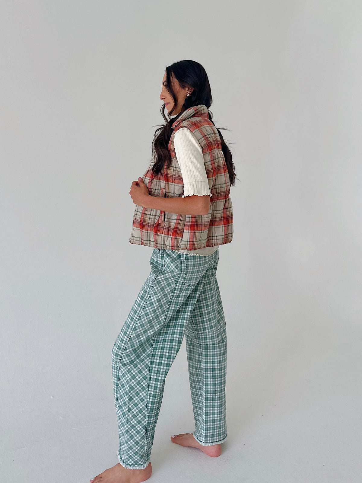 A woman stands barefoot in profile wearing a sleeveless red and beige plaid vest over a white short-sleeve top and green-and-white plaid pants, against a plain light gray studio background.