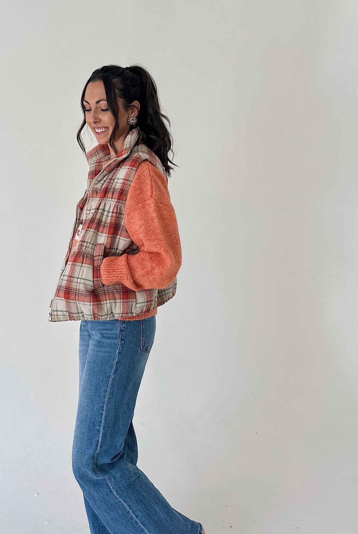 A woman wearing a sleeveless plaid vest with orange knit sleeves and blue jeans, standing against a plain off-white wall.