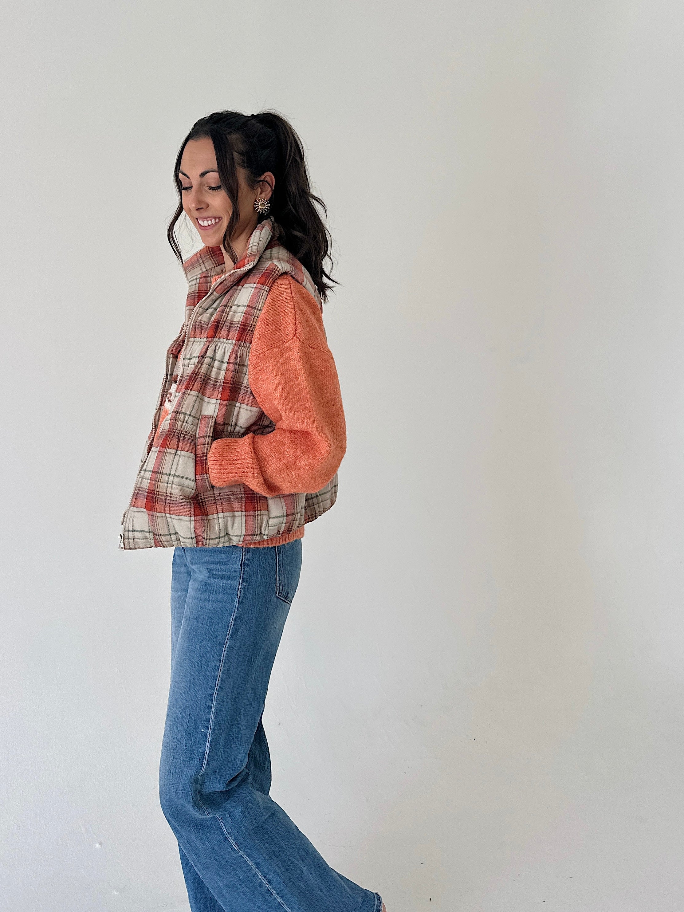 A woman wearing a sleeveless plaid vest with orange knit sleeves and blue jeans, standing against a plain off-white wall.