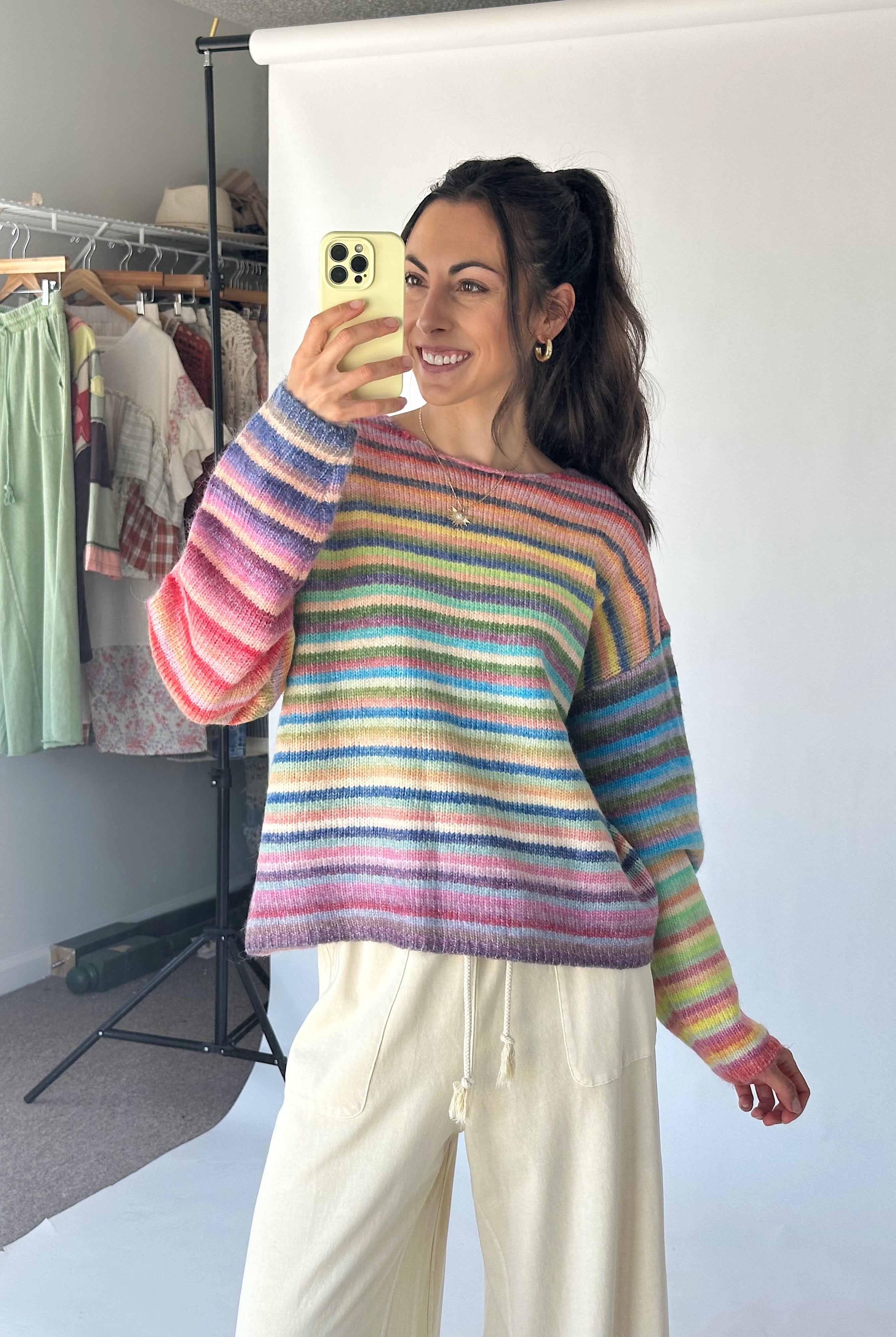 A woman takes a mirror selfie wearing a pastel rainbow striped knit sweater and cream drawstring pants in a studio setting with a clothing rack in the background.