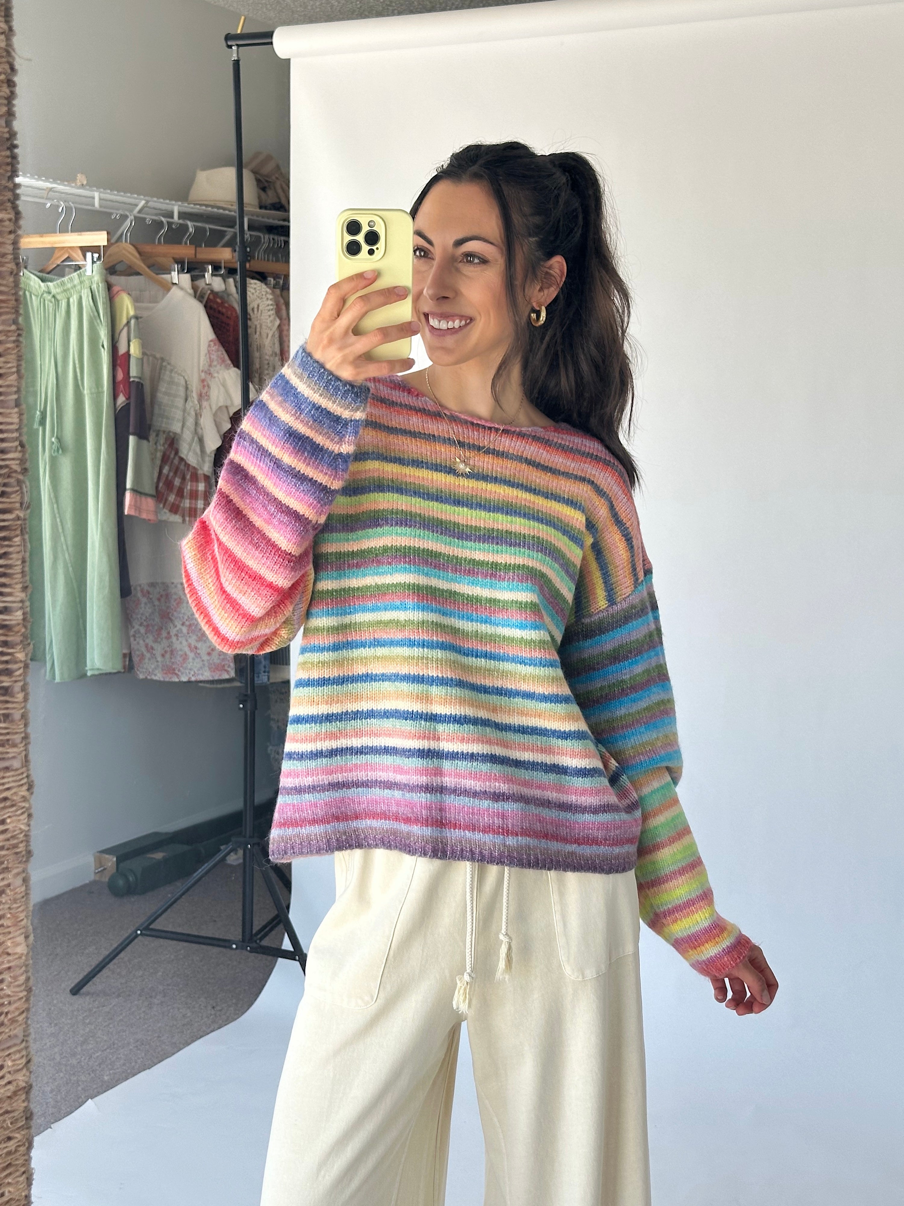 A woman takes a mirror selfie wearing a pastel rainbow striped knit sweater and cream drawstring pants in a studio setting with a clothing rack in the background.