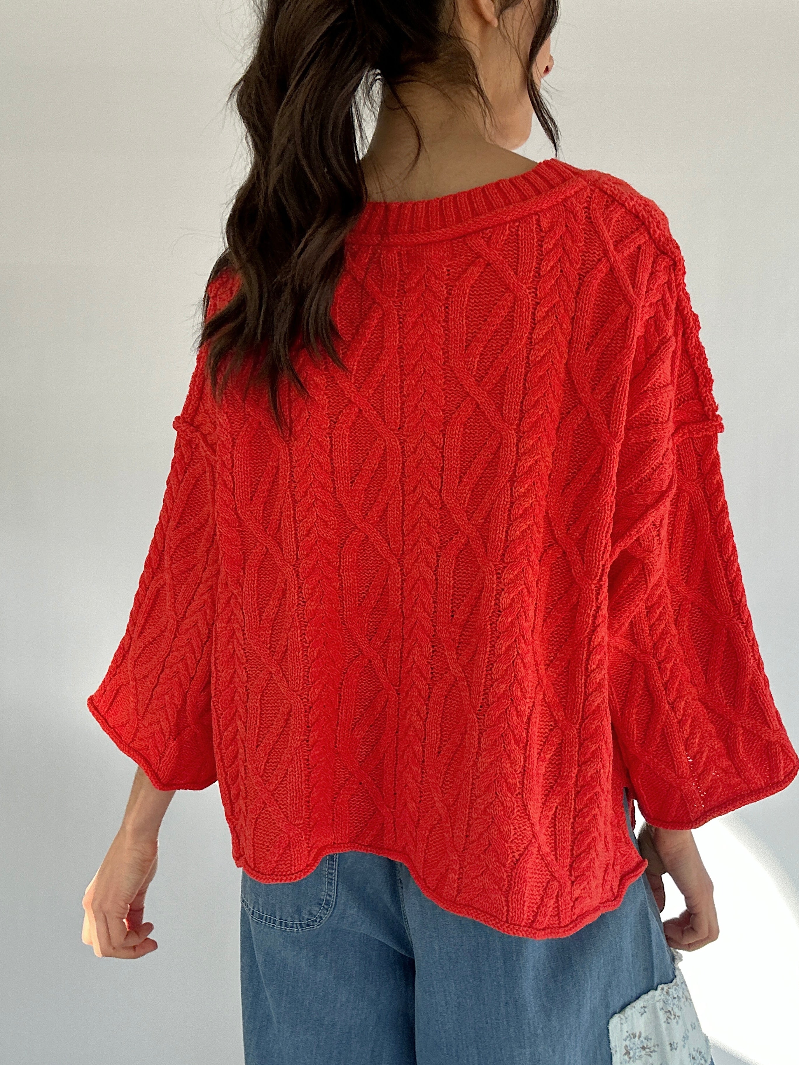 Person wearing a bright coral cable knit sweater with a plain background showing the back
