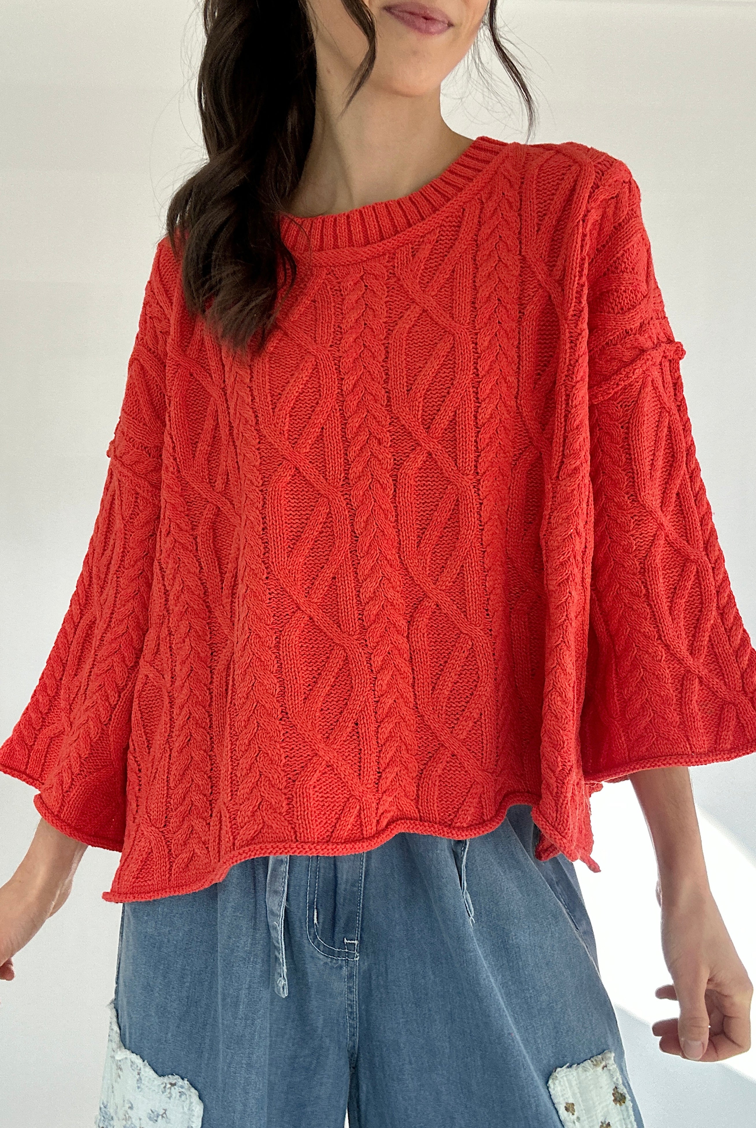 Person wearing a bright coral cable knit sweater and blue jeans on a white background