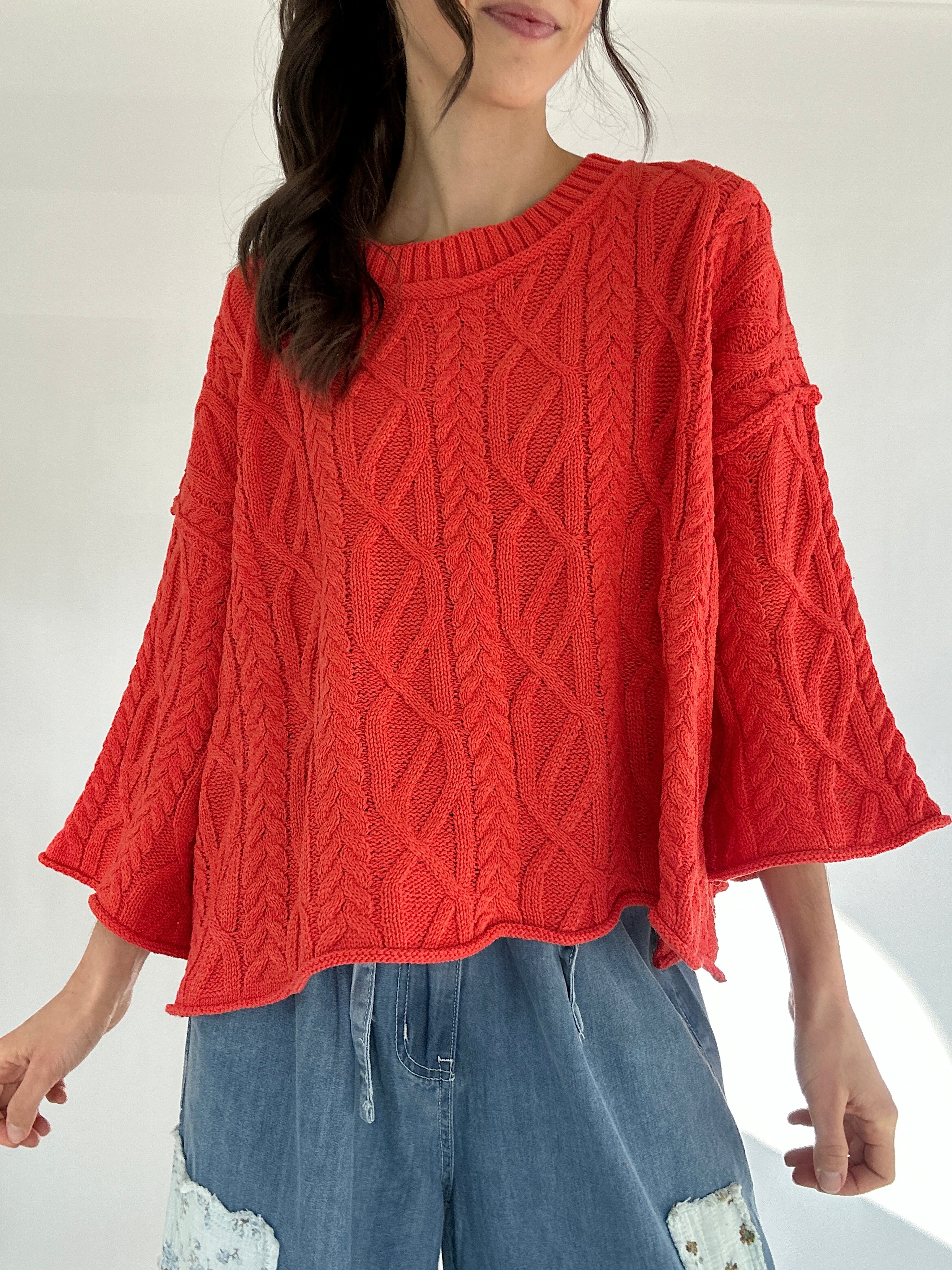 Person wearing a bright coral cable knit sweater and blue jeans on a white background