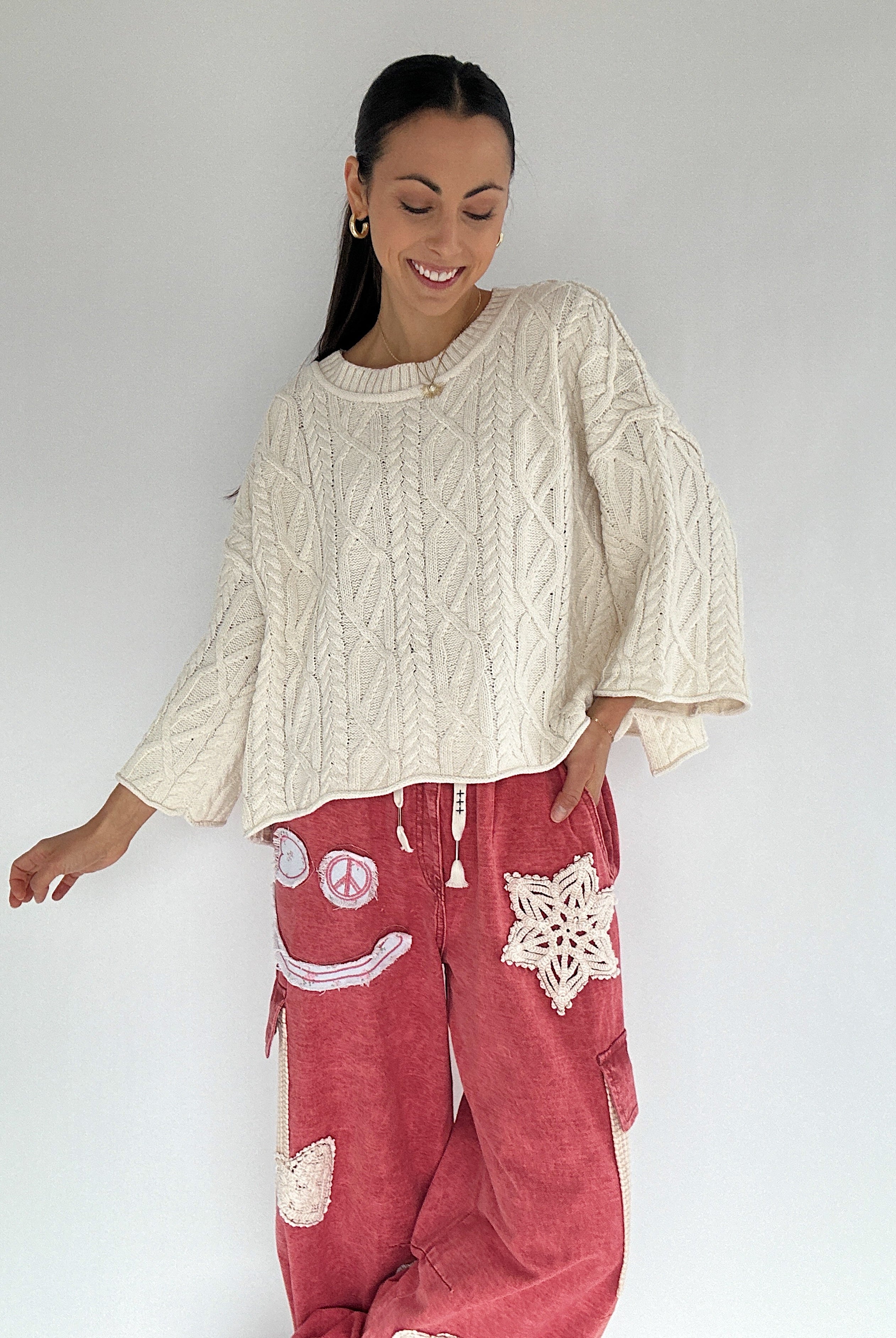 Woman wearing a cream cable knit sweater and red pants with patches on a white background