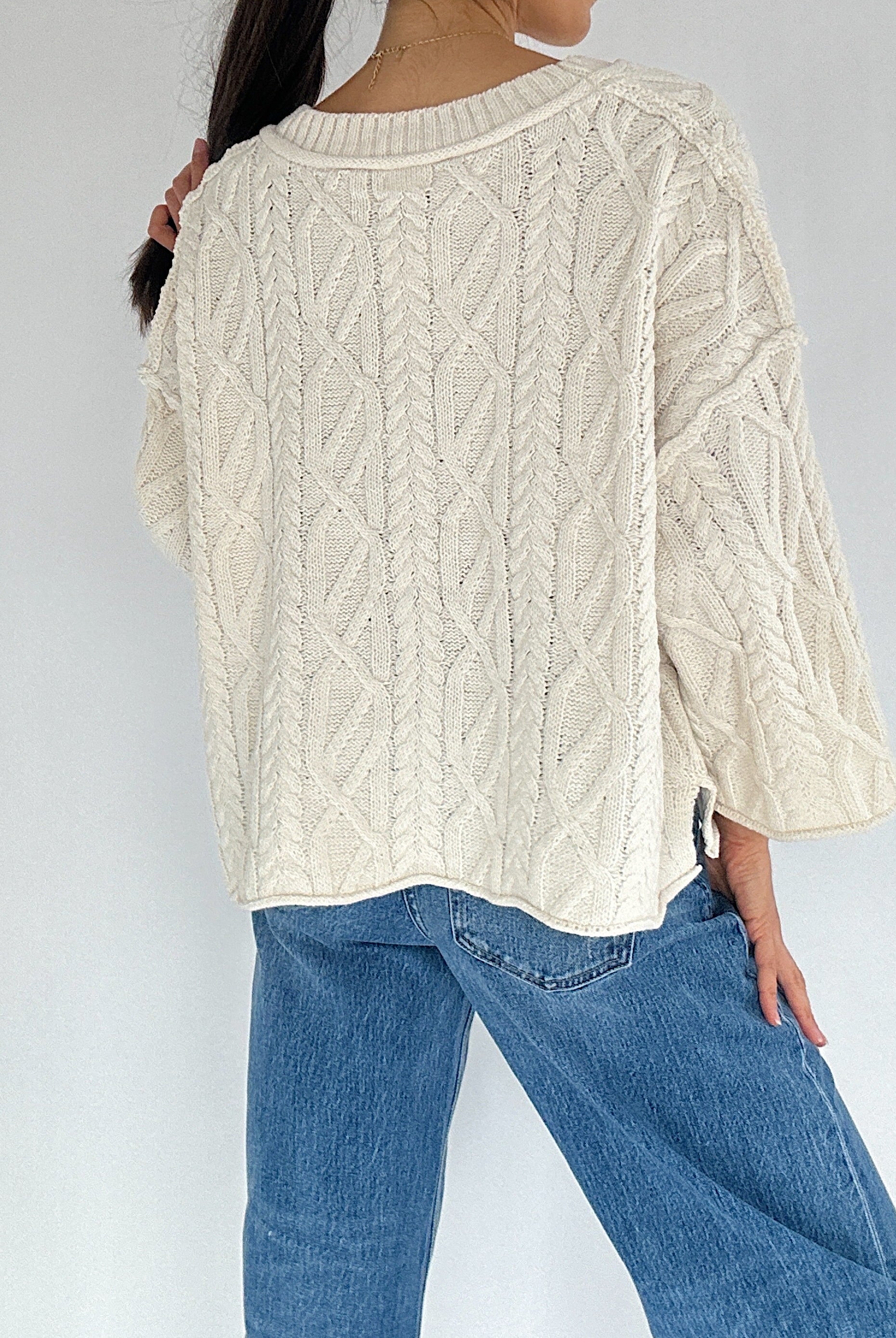 Person wearing a cream cable knit sweater and blue jeans against a white background back view