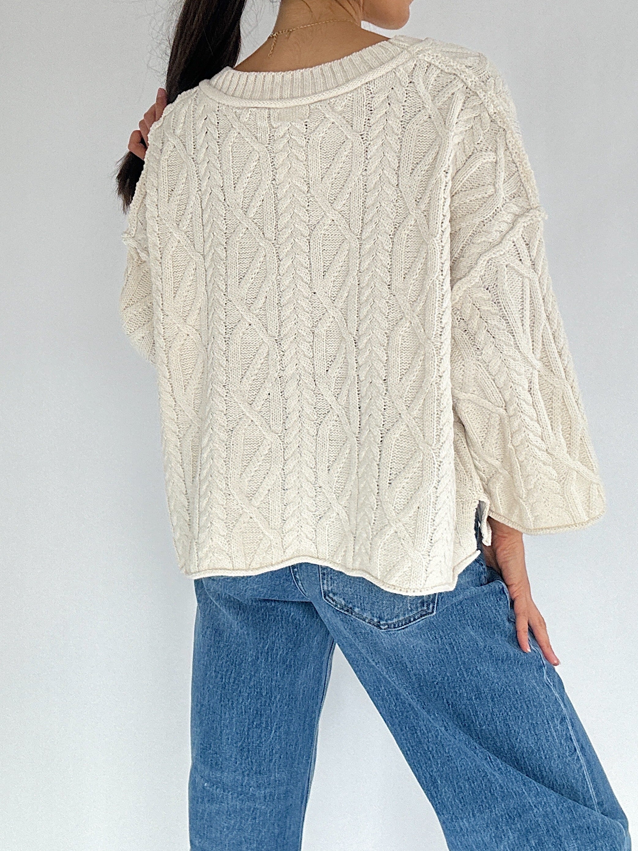 Person wearing a cream cable knit sweater and blue jeans against a white background back view