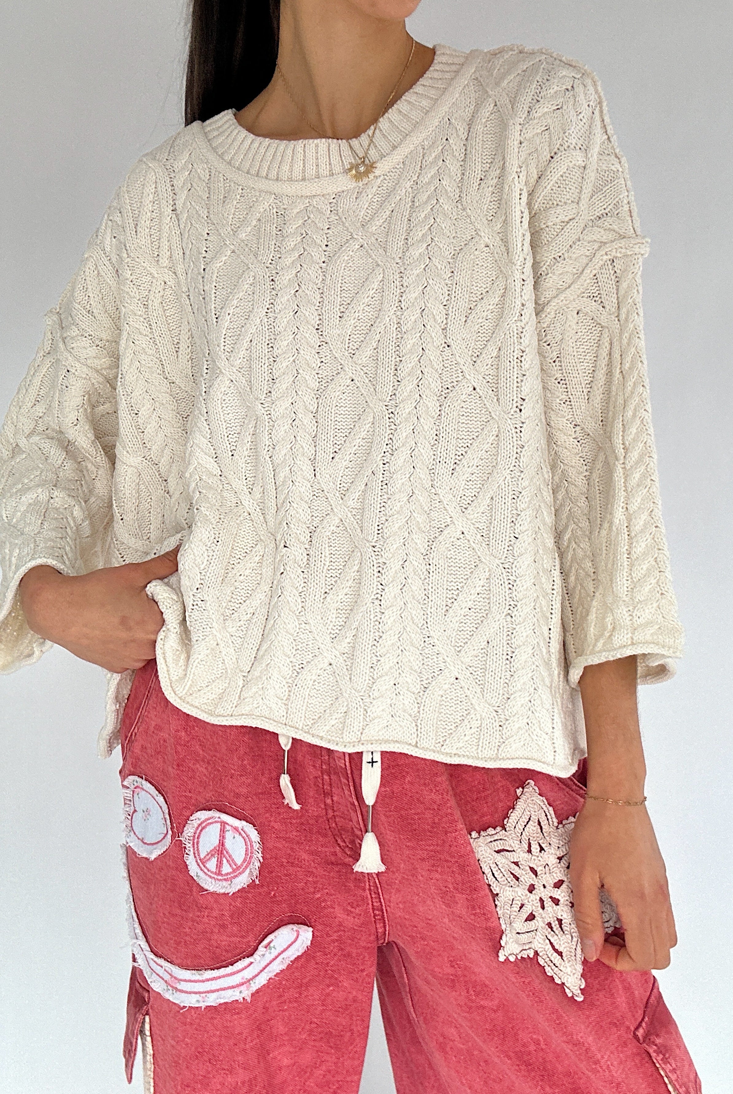 Person wearing a cream cable knit sweater and red pants with smiley face patch design.