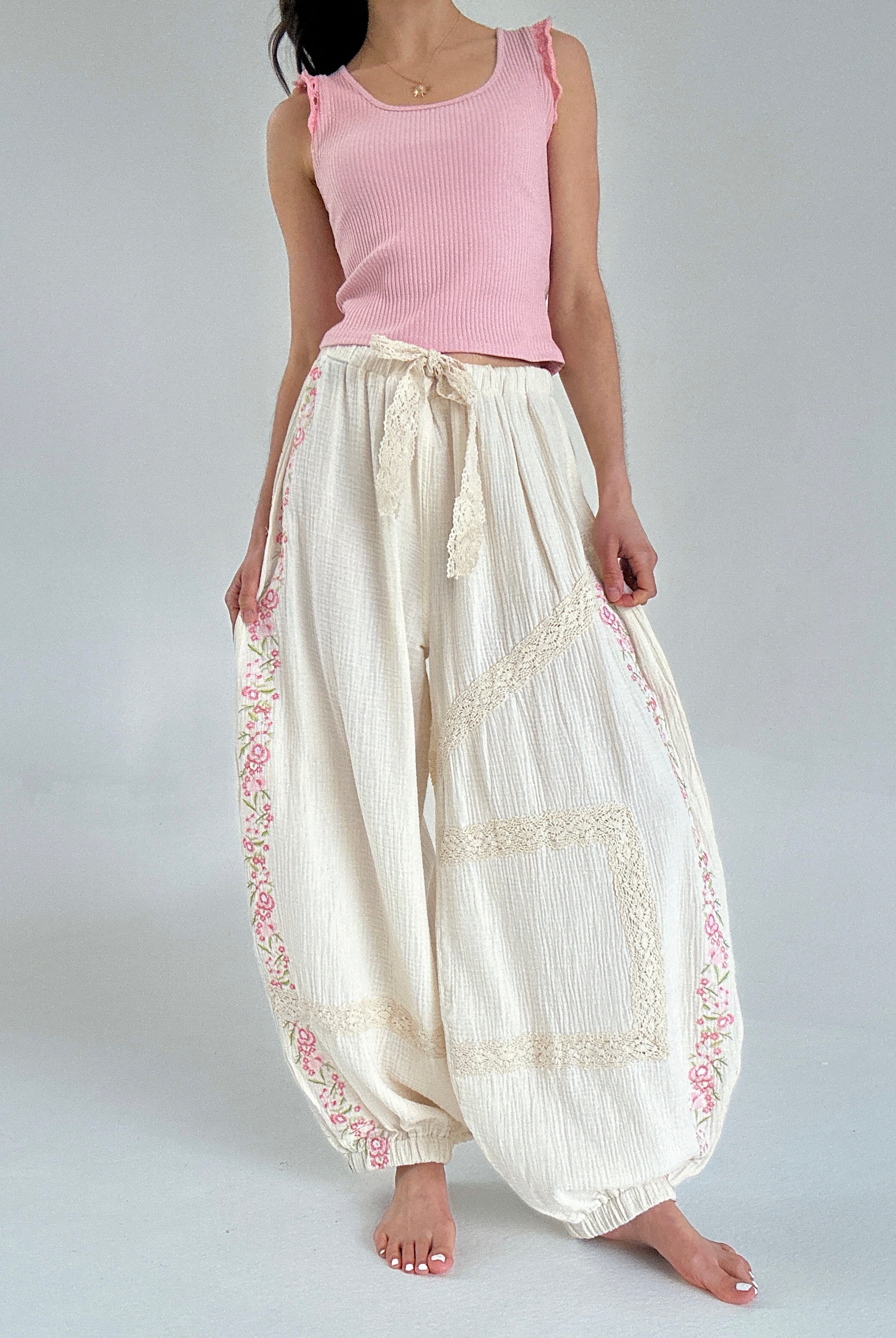 Person wearing a pink sleeveless top and white pants with lace and embroidery details on a plain background