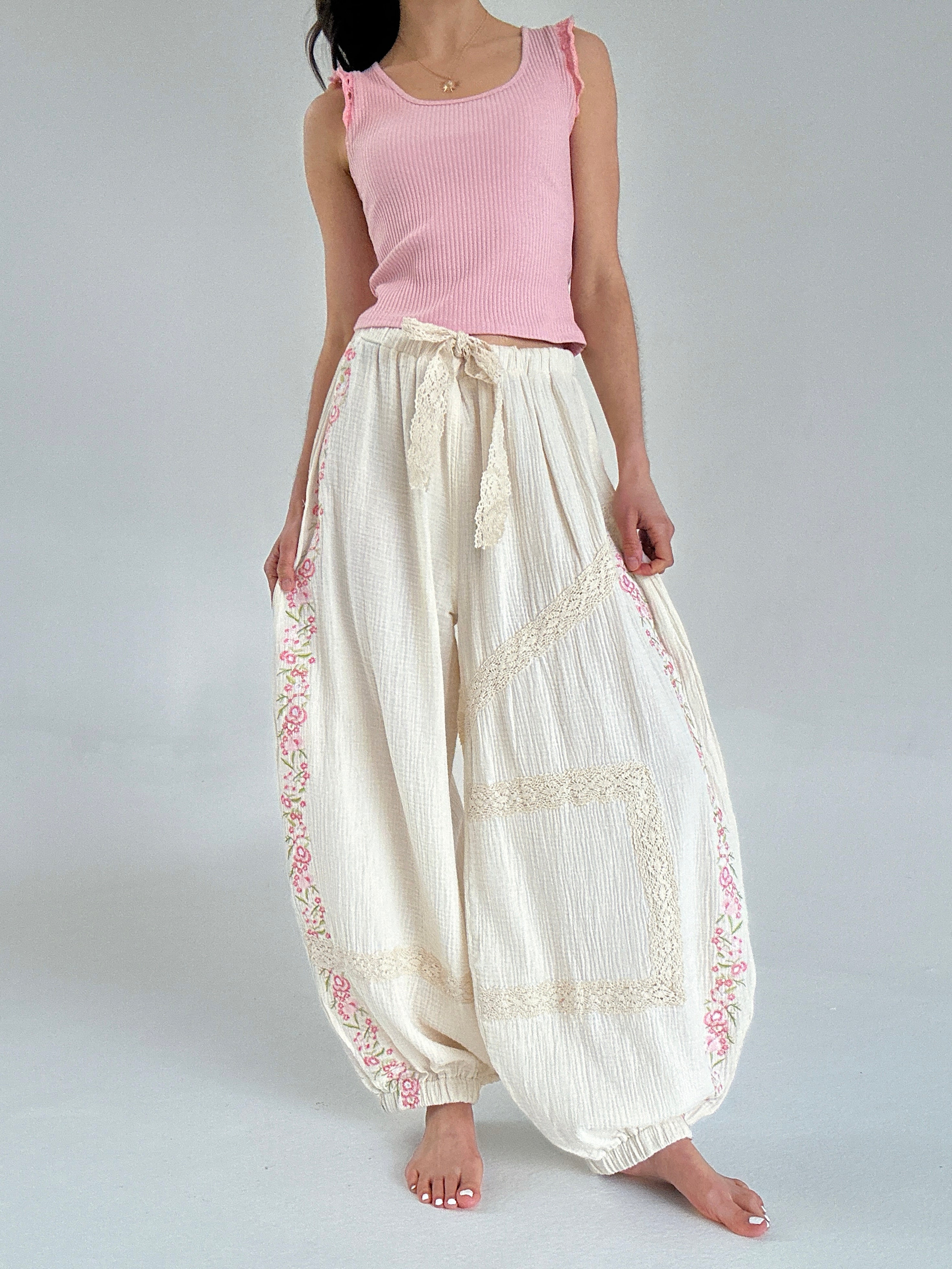 Person wearing a pink sleeveless top and white pants with lace and embroidery details on a plain background