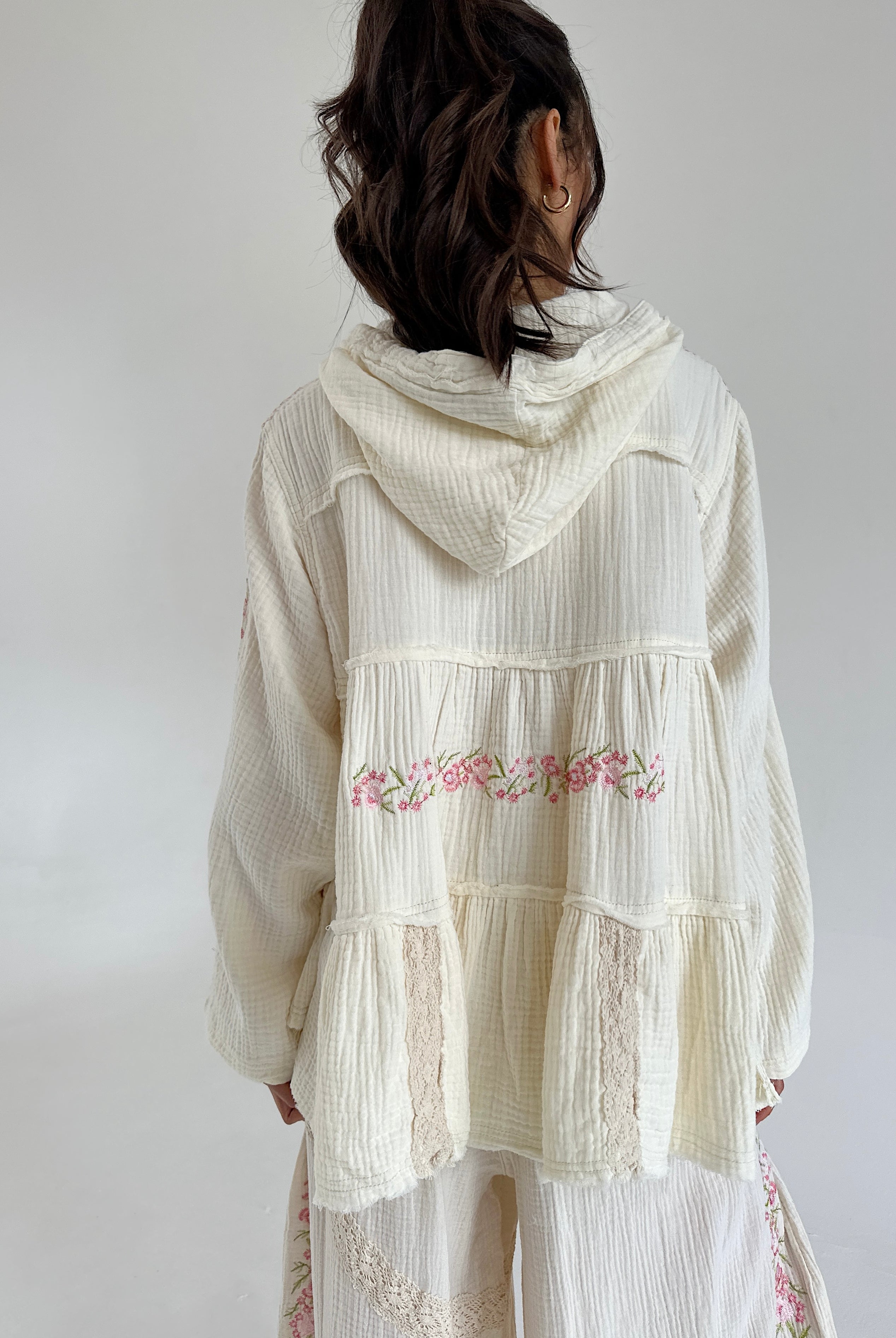 Woman wearing a white embroidered hooded top with a plain background showing the back