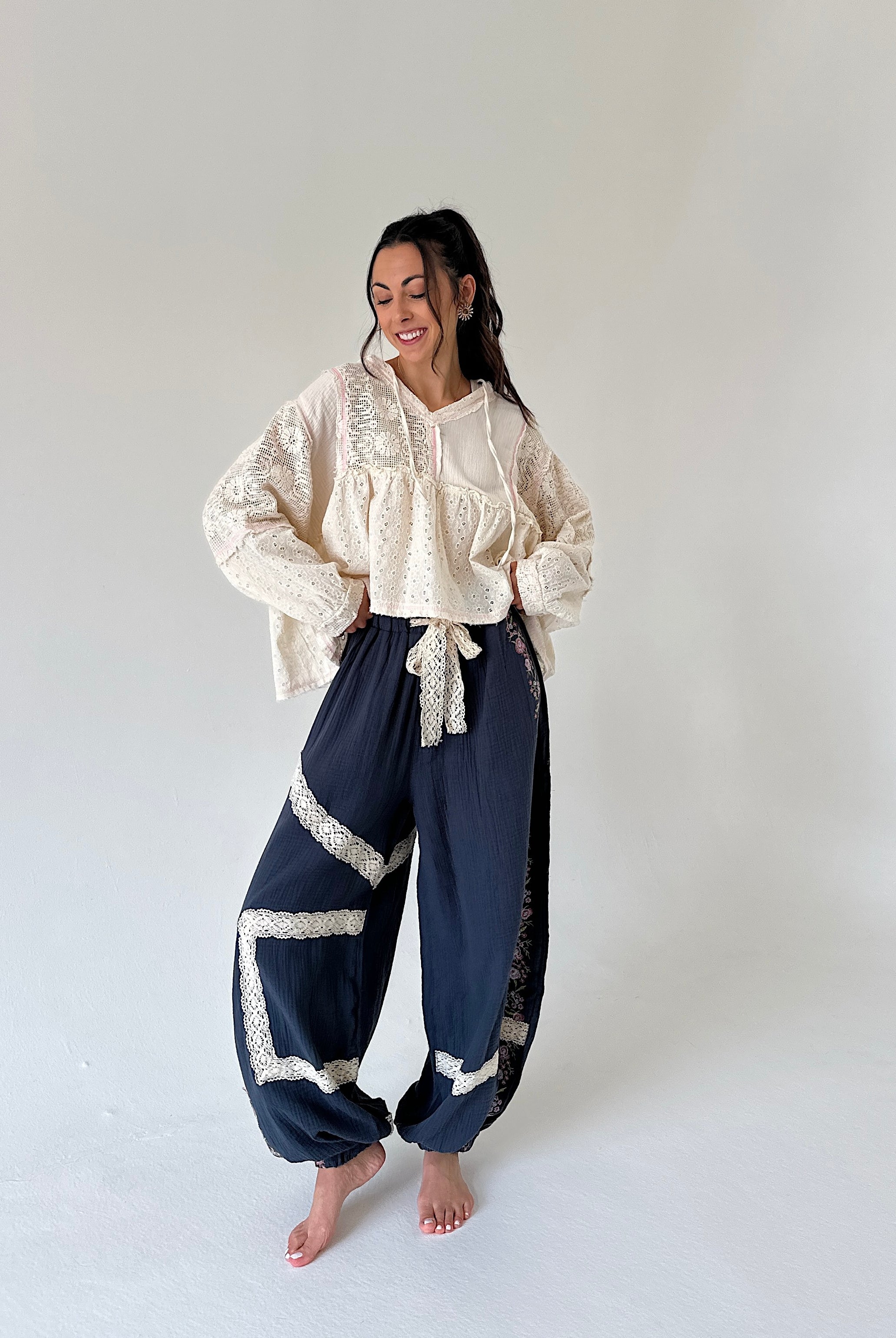 A woman stands barefoot in a studio wearing a cream lace top and matching with dark navy wide-leg pants featuring cream lace trim.