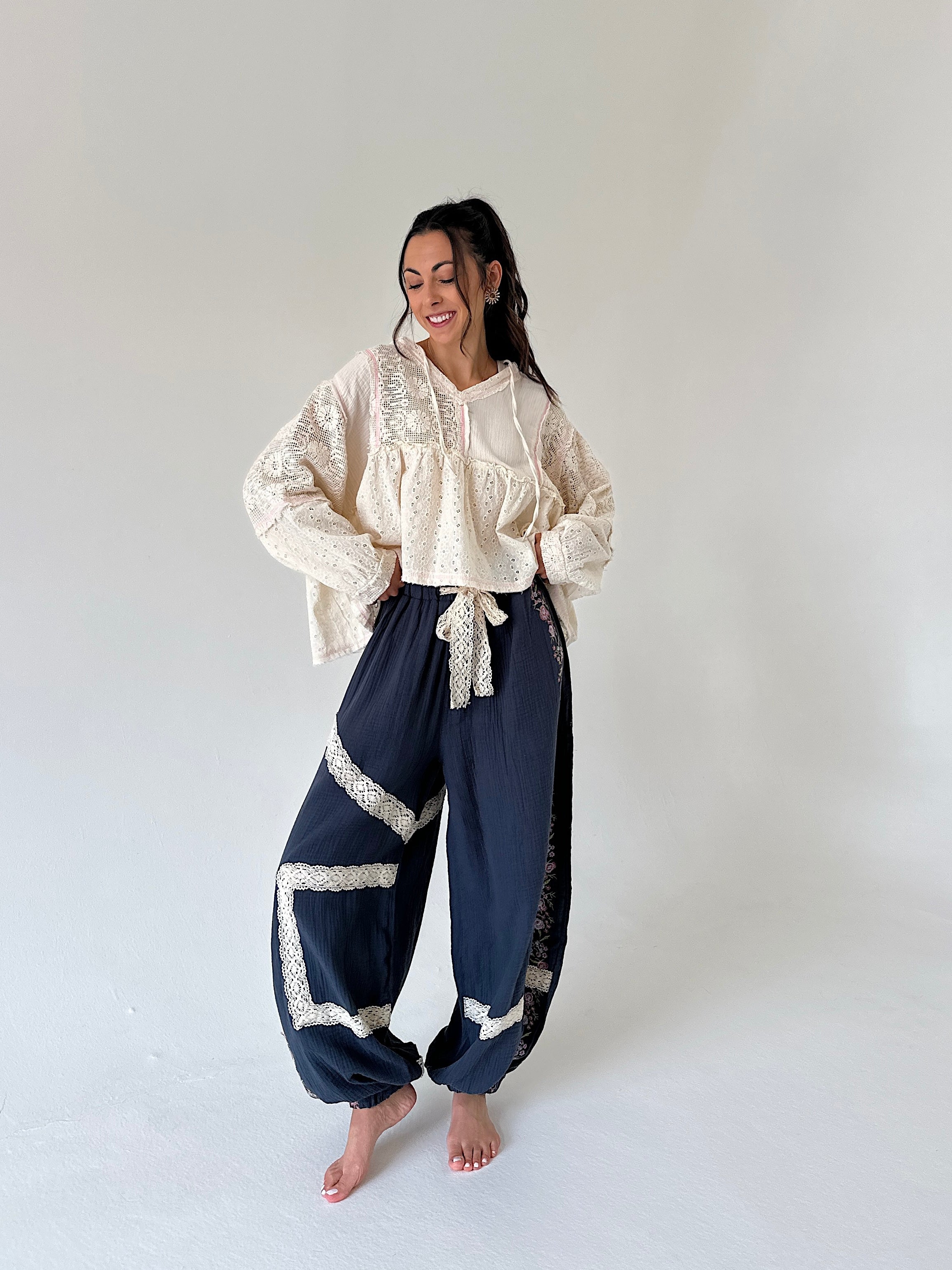 A woman stands barefoot in a studio wearing a cream lace top and matching with dark navy wide-leg pants featuring cream lace trim.