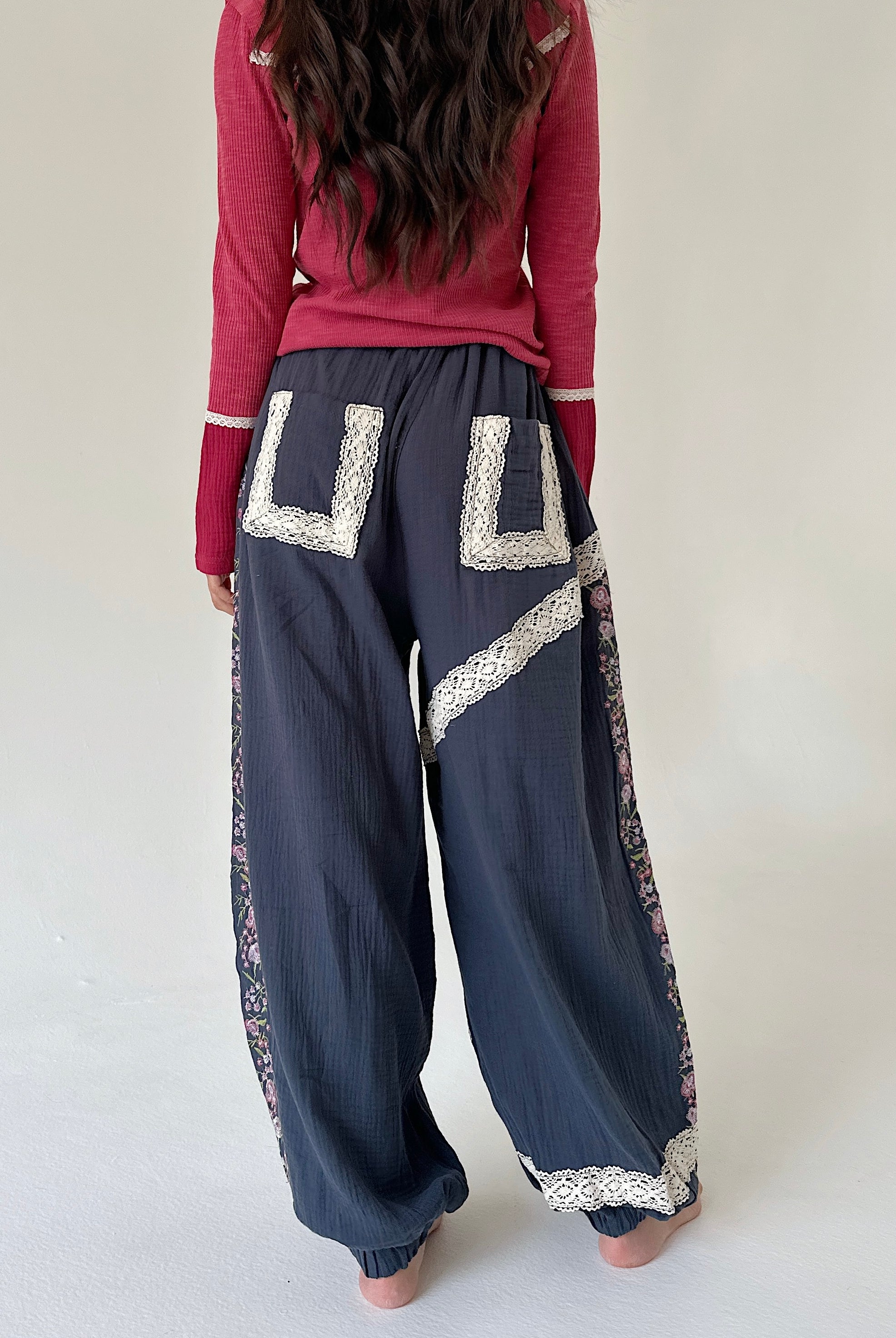 A person shown from the back wearing loose dark blue pants with large cream lace applique details and a pink long-sleeve top, standing against a plain light background.