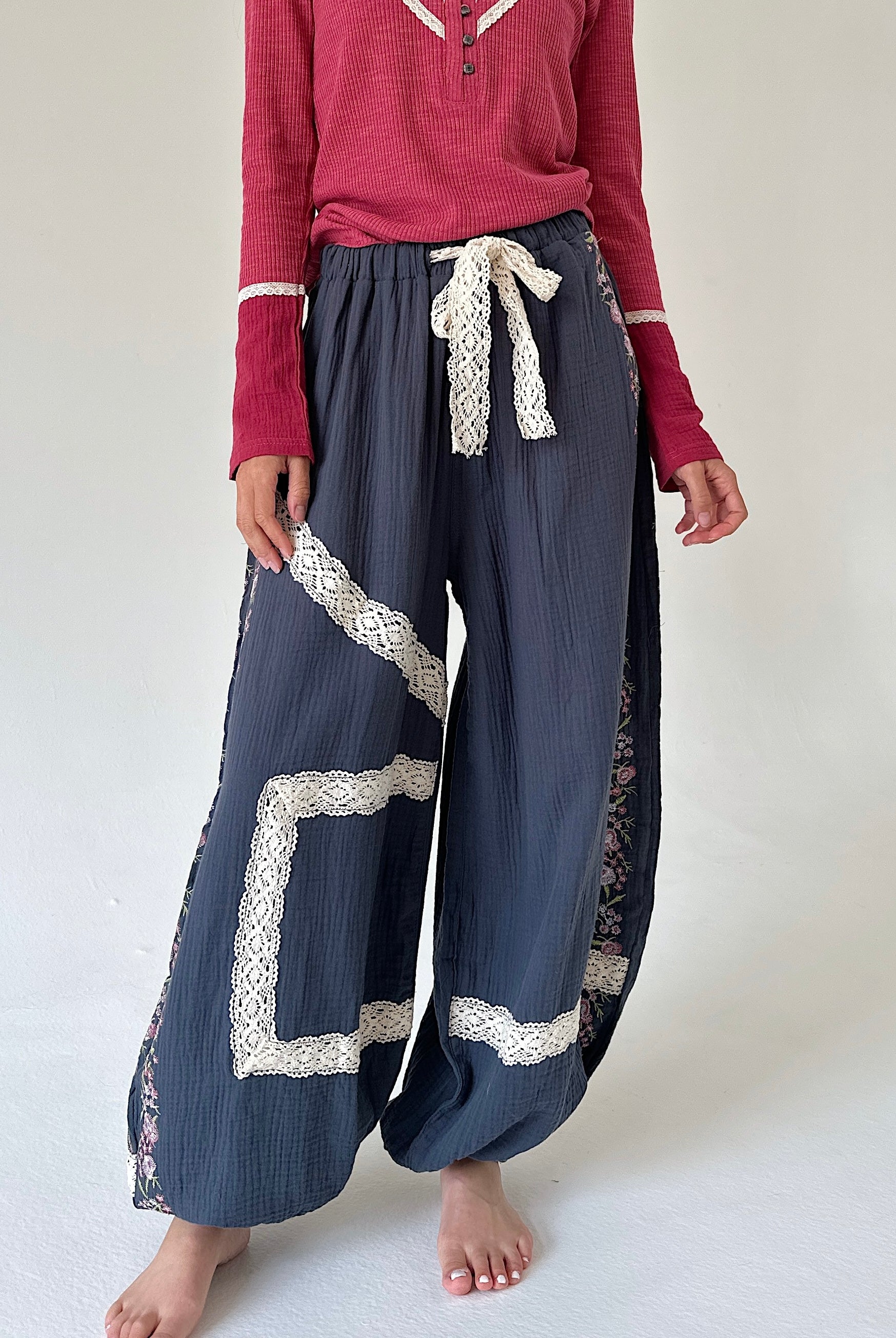 A person wearing loose, wide-leg navy pants with cream lace trim and a matching cream lace tie at the waist, paired with a burgundy long-sleeve henley top. The photo is cropped from the chest down against a plain off-white studio background.