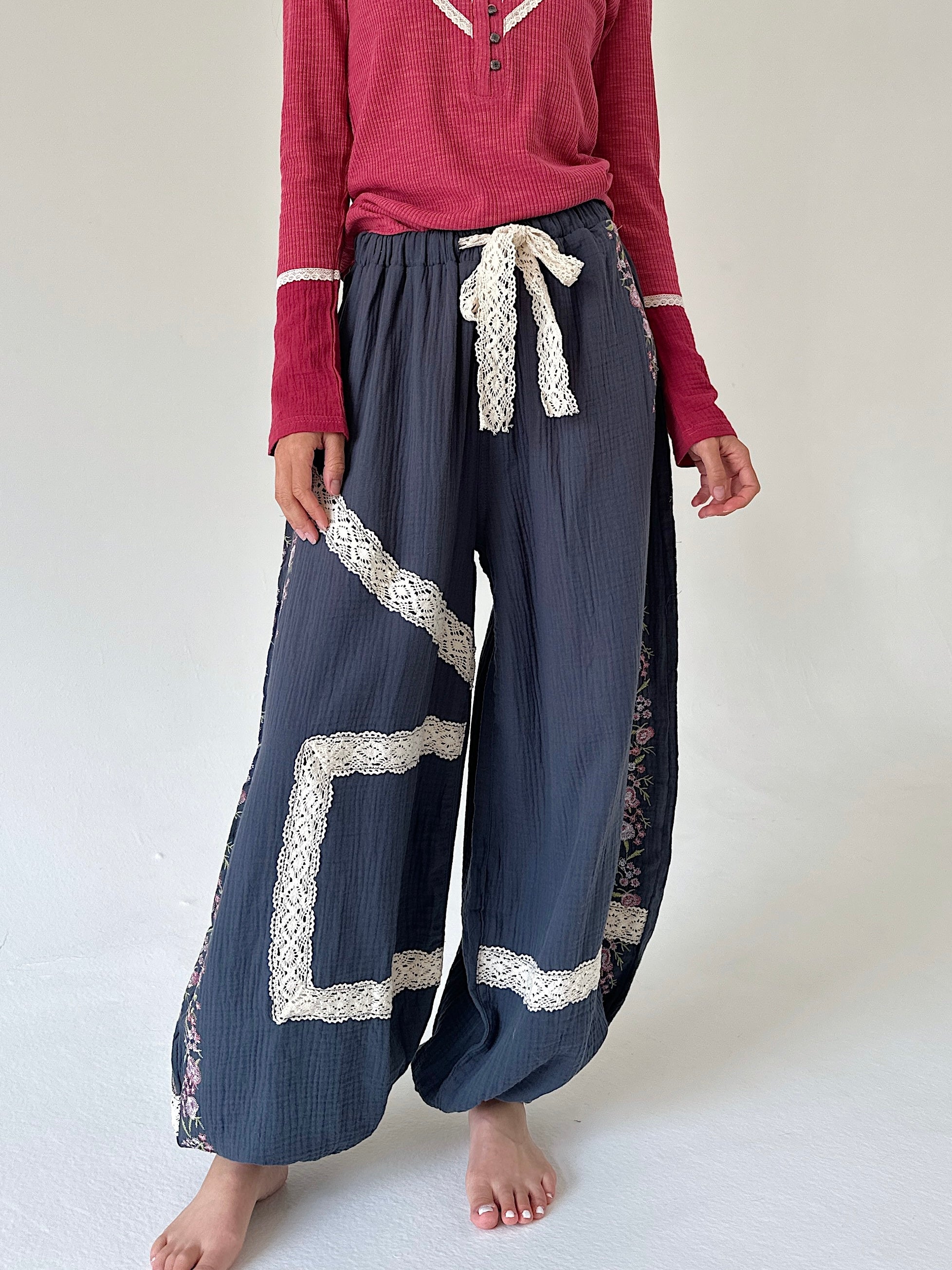A person wearing loose, wide-leg navy pants with cream lace trim and a matching cream lace tie at the waist, paired with a burgundy long-sleeve henley top. The photo is cropped from the chest down against a plain off-white studio background.