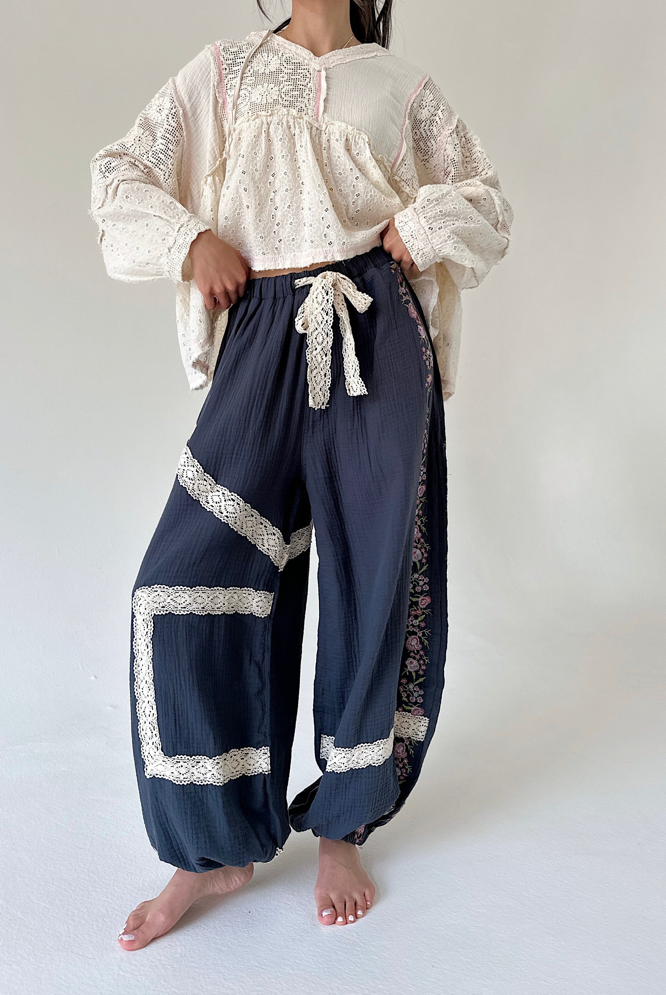A model wearing a cream lace-trimmed long-sleeve blouse and navy blue gauze harem-style pants with white lace trim and a white drawstring tie, standing barefoot against a plain off-white studio background.